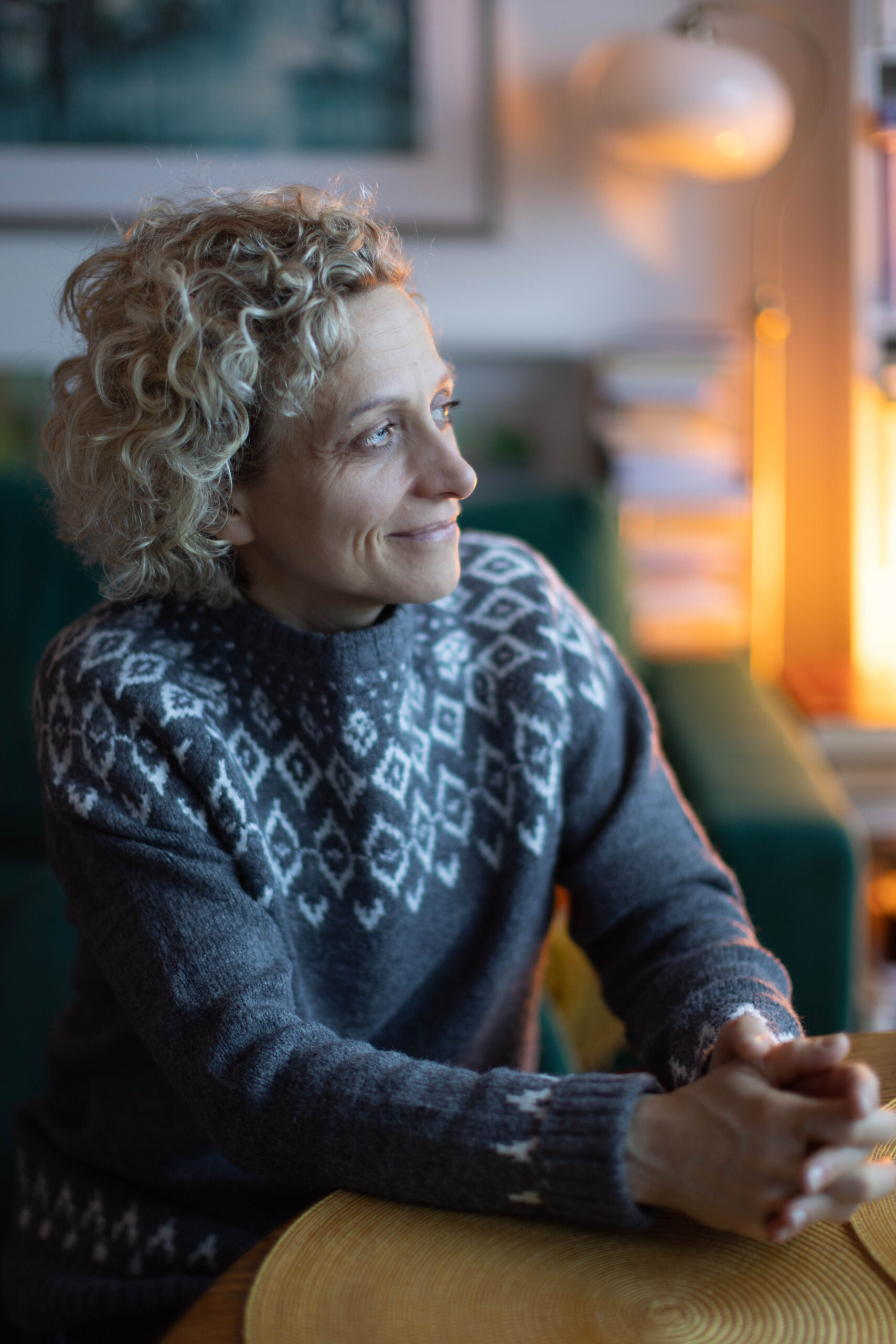 18. Lekko uśmiechnięta zamyślona kobieta o jasnych kręconych włosach przy stole patrząca w okno