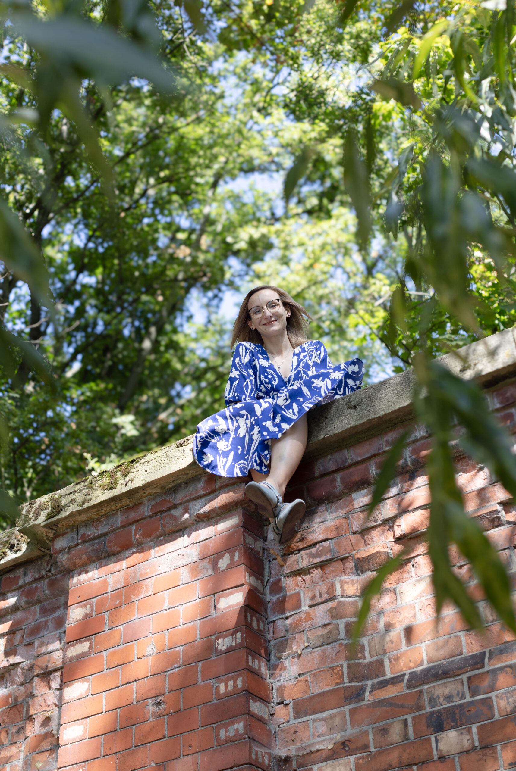 młoda kobieta blondynka w biało-niebieskiej sukience z uśmiechem pozuje do zdjęcia siedząc na akwedukcie warszawskiej cytadeli