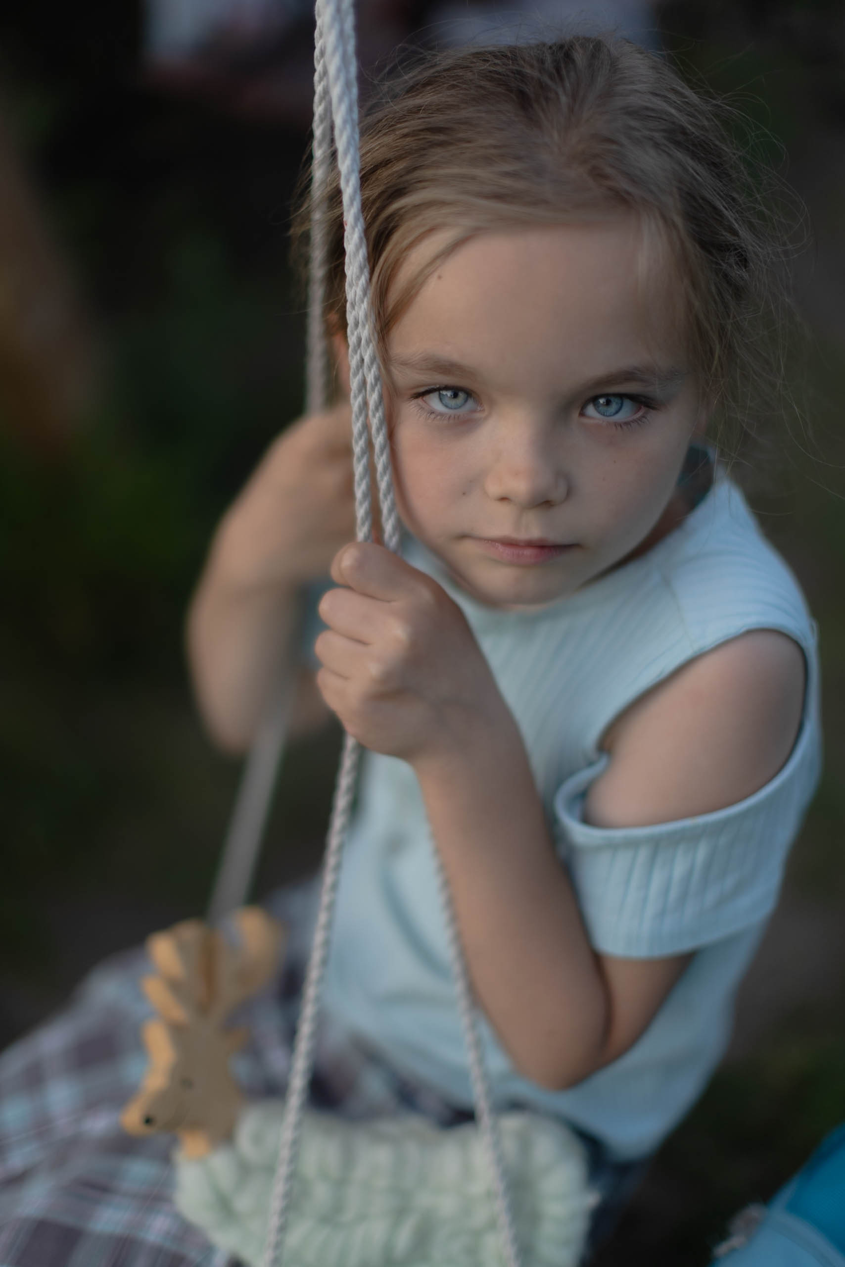 dziewczynka 9-letnia o niebieskich oczach, jasnych włosach i w niebieskiej bluzeczce na huśtawce, patrzy prosto w obiektyw