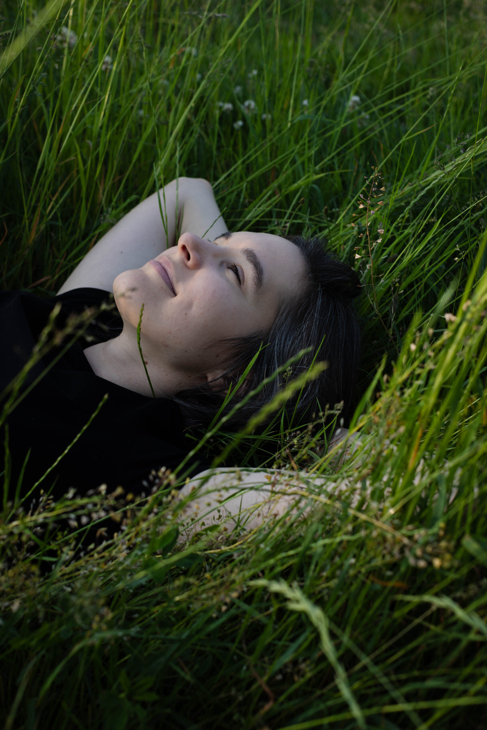 Młoda ładna brunetka leży w trawie z uśmiechem, przymkniętymi oczami i rękami założonymi za głowę