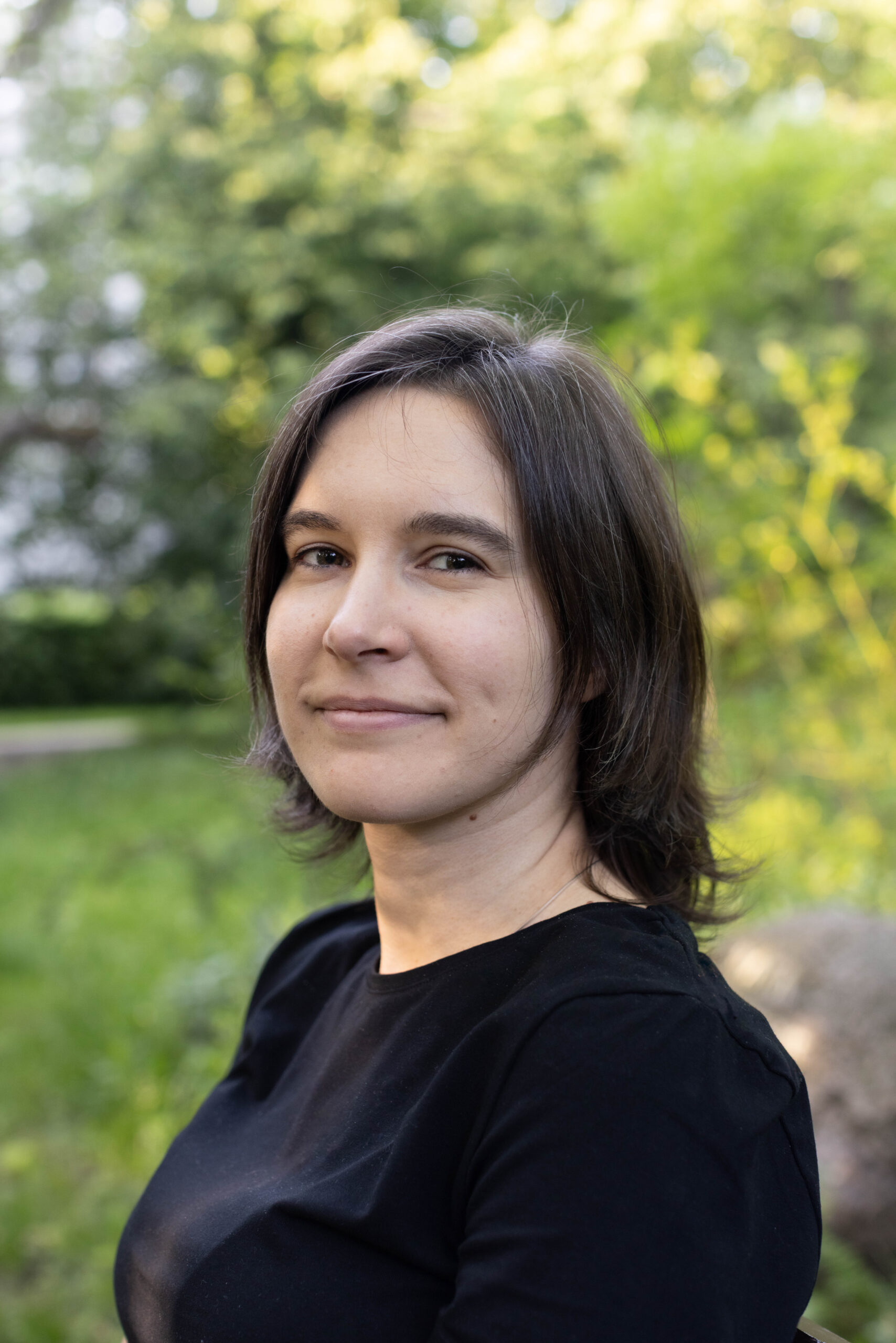 Młoda ładna brunetka - typ kolorystyczny Zima - w czarnym podkoszulku, z półuśmiechem pozuje do zdjęcia w parku wśród zieleni