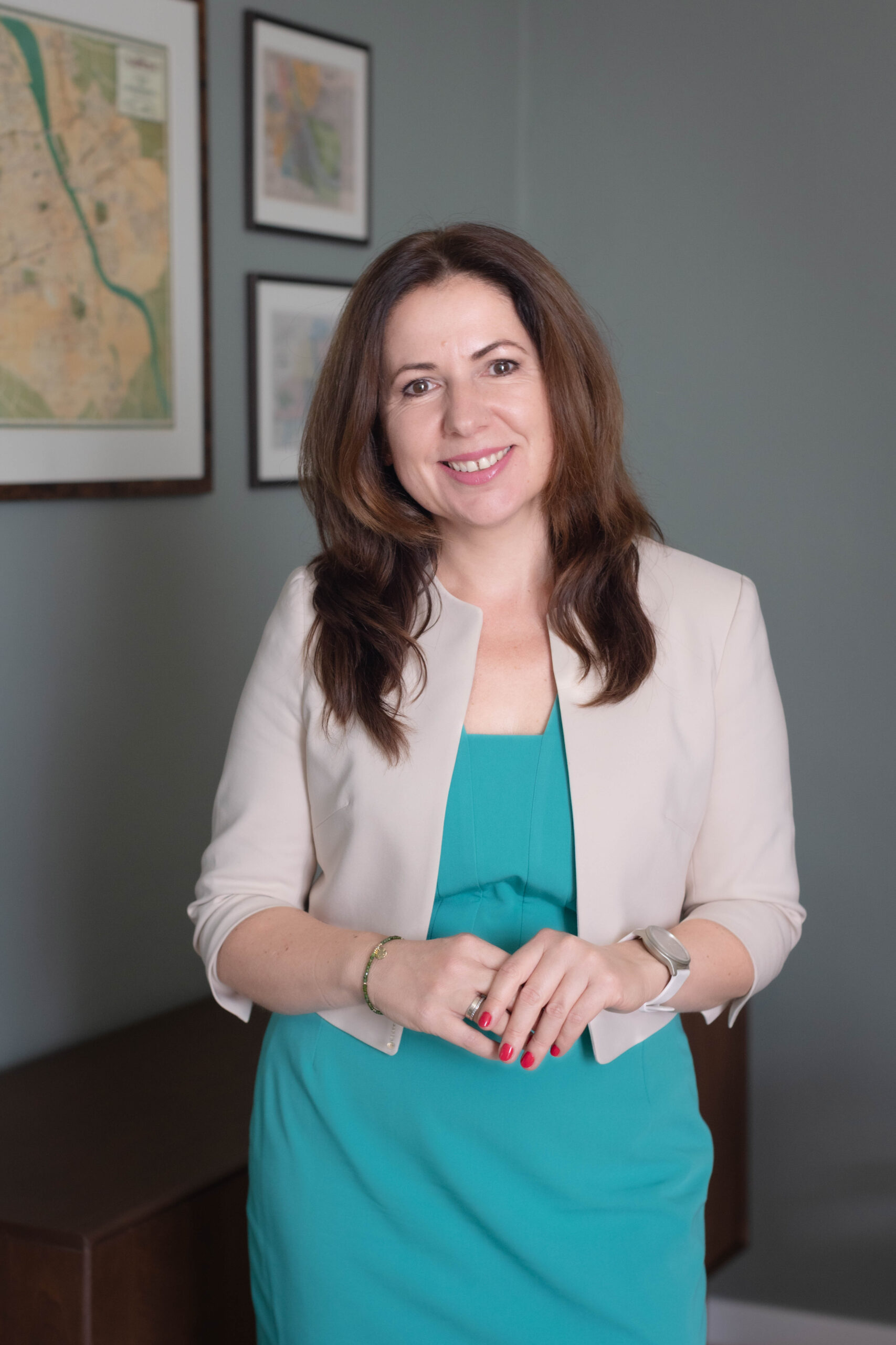 Uśmiechnięta kobieta szatynka w eleganckiej turkusowej sukience i kremowej marynarce, która pozuje do zdjęcia wizerunkowego w swojej kancelarii prawnej