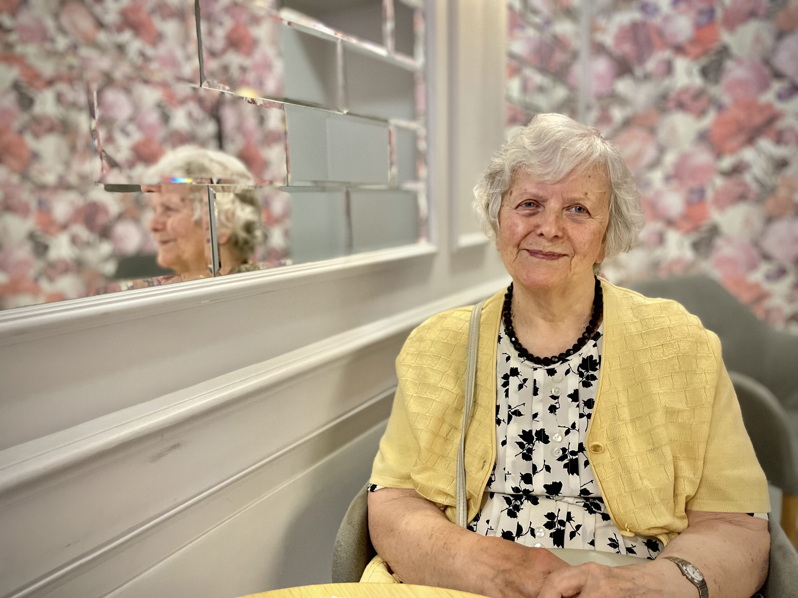 Starsza kobieta o siwych kręconych włosach kolorowo ubrana siedzi w kawiarni przy stoliku na tle ściany w kwiecistą tapetę i odbija się w lustrze