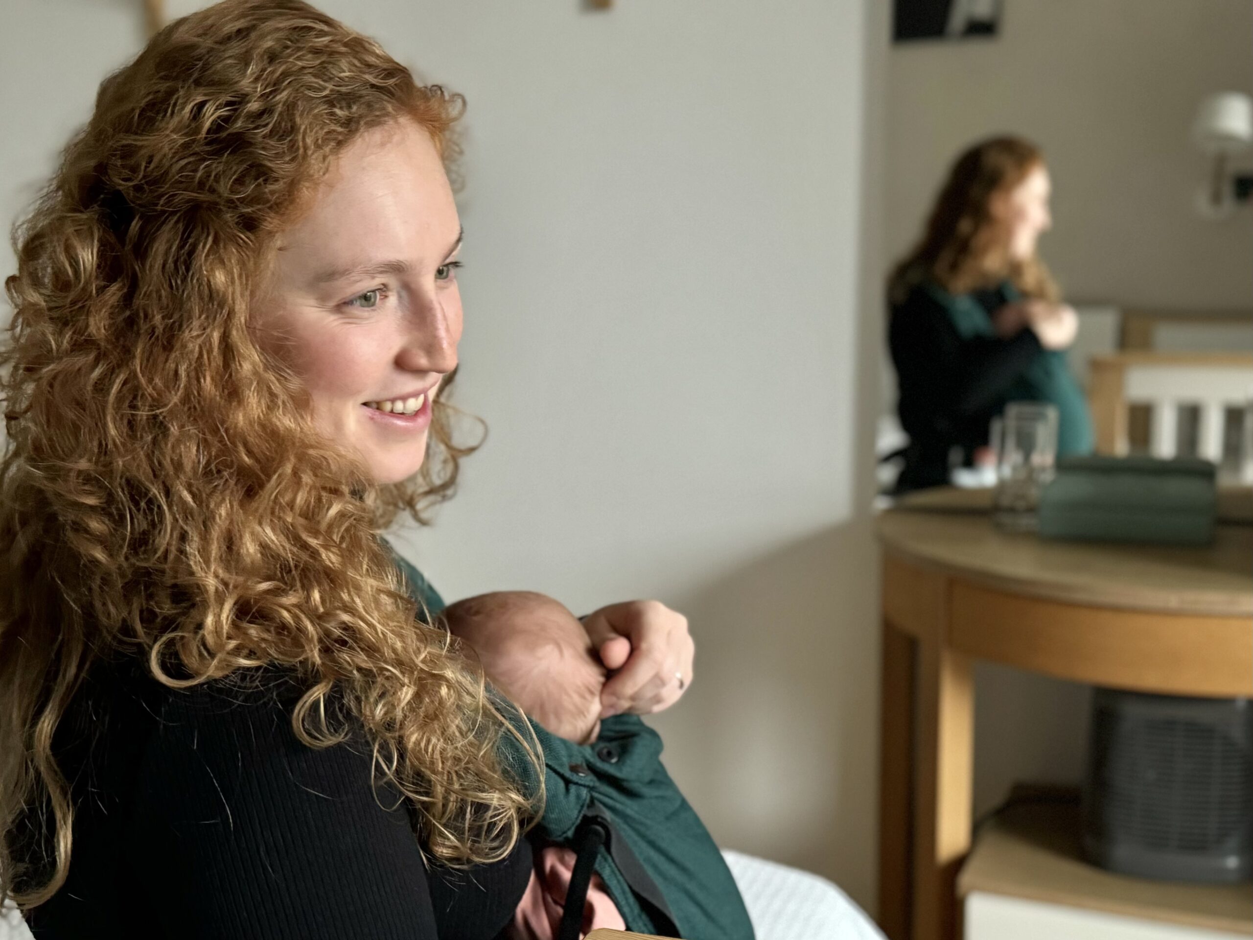 Młoda matka o rusych kręconych włosach z uśmiechem tuli niemowlę odbijając się w lustrze podczas sesji zdjęciowej