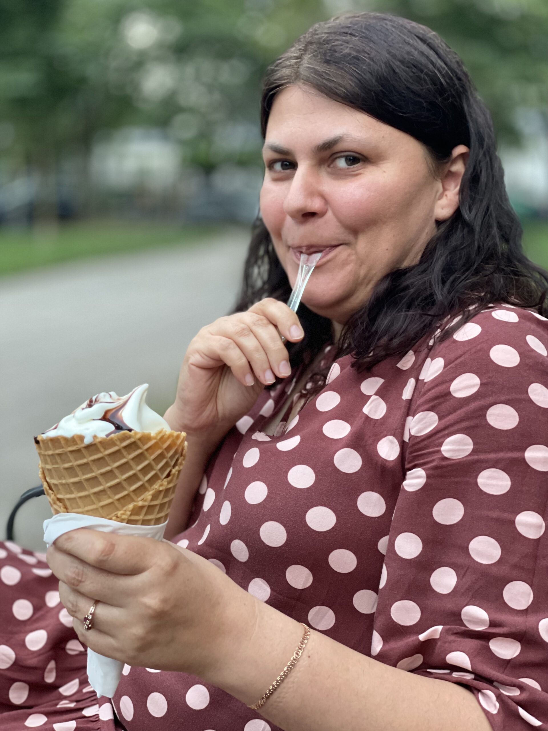 Kobieta brunetka w brązowej sukni w grochy, lekko tęga, która trzyma w dłoni loda i z wielkim apetytem go degustuje