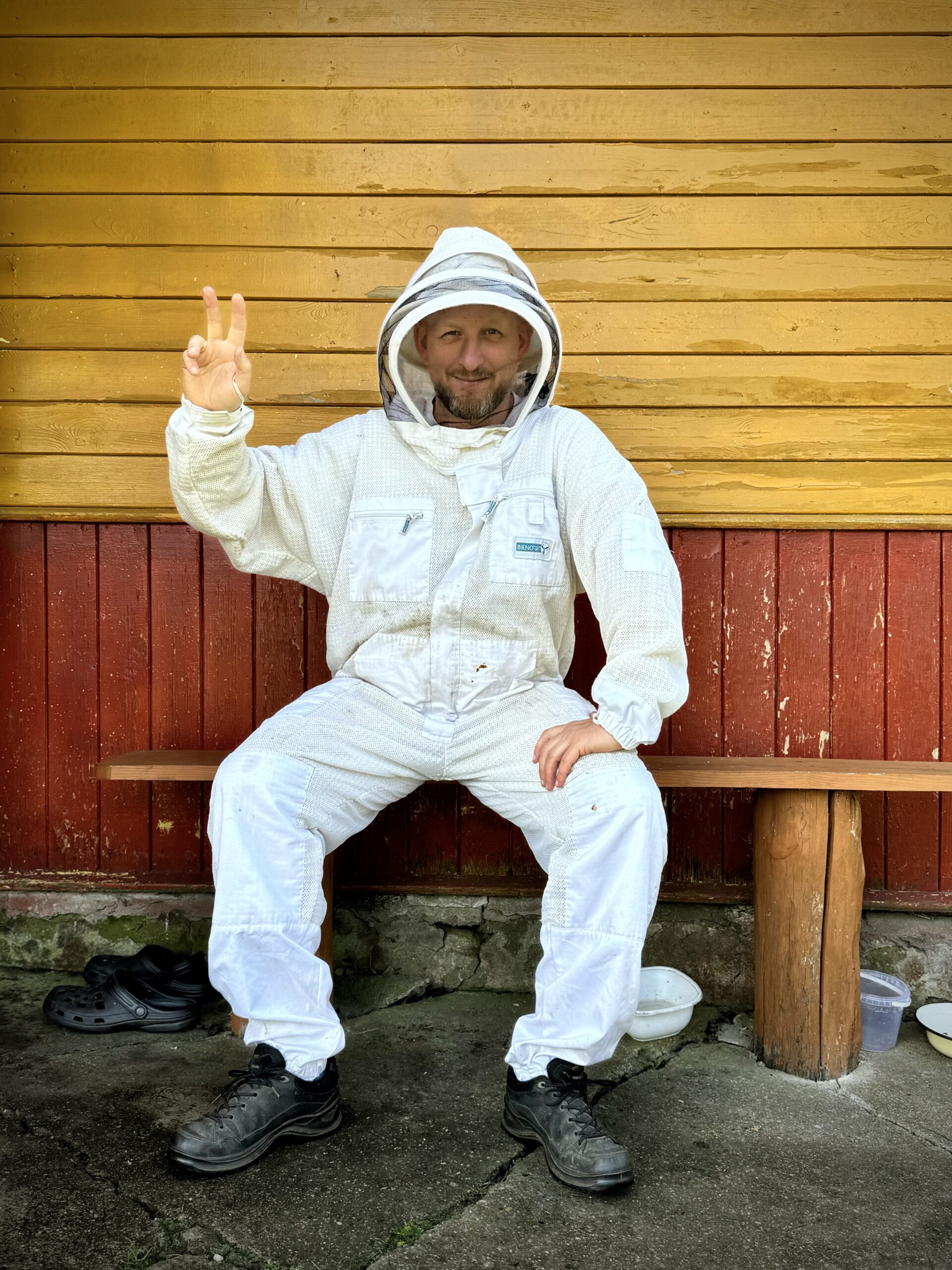 pszczelarz w specjalnym stroju ochronnym siedzący na ławce z uśmiechem i pokazujący palcami gest zwycięstwa