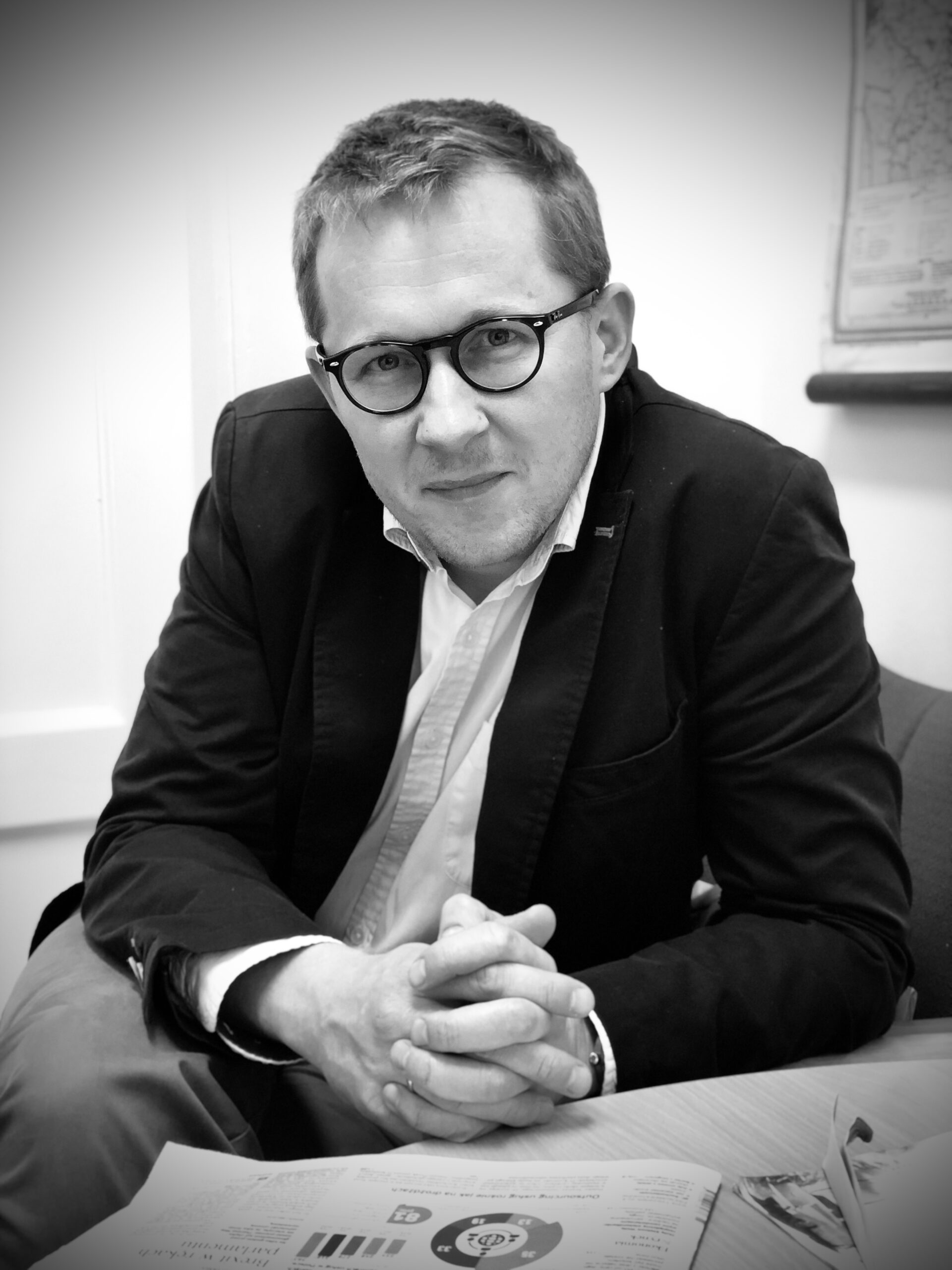 Czarno-biały portret mężczyzny około czterdziestki, w koszuli i marynarce, siedzącego bokiem przy biurku, nad gazetą i z uśmiechem patrzącego w obiektyw aparatu