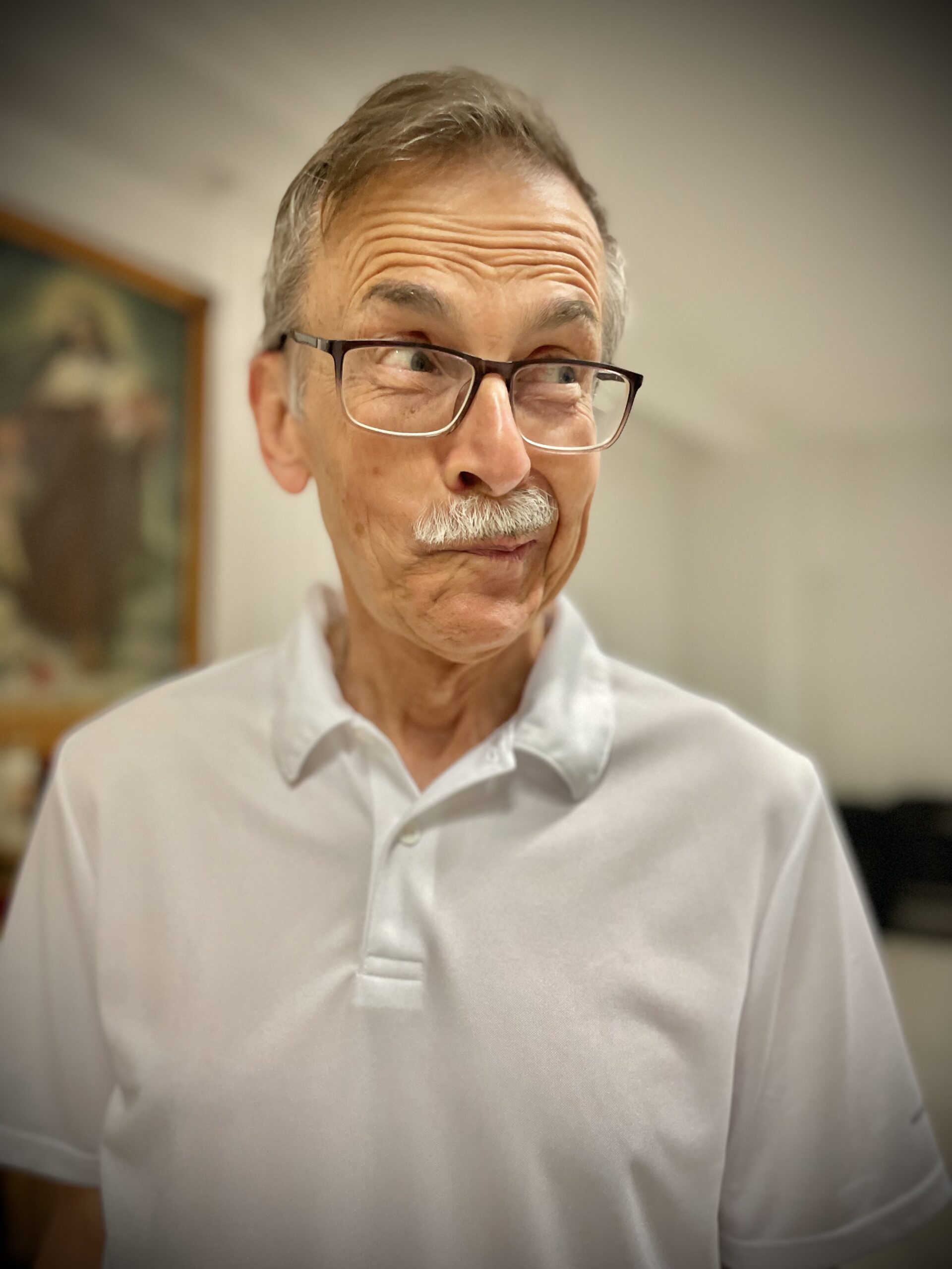 Portret starszego mężczyzny z siwym wąsikiem i w okularach, w białej koszulce polo, który wygłupia się do zdjęcia i robi zabawną minę