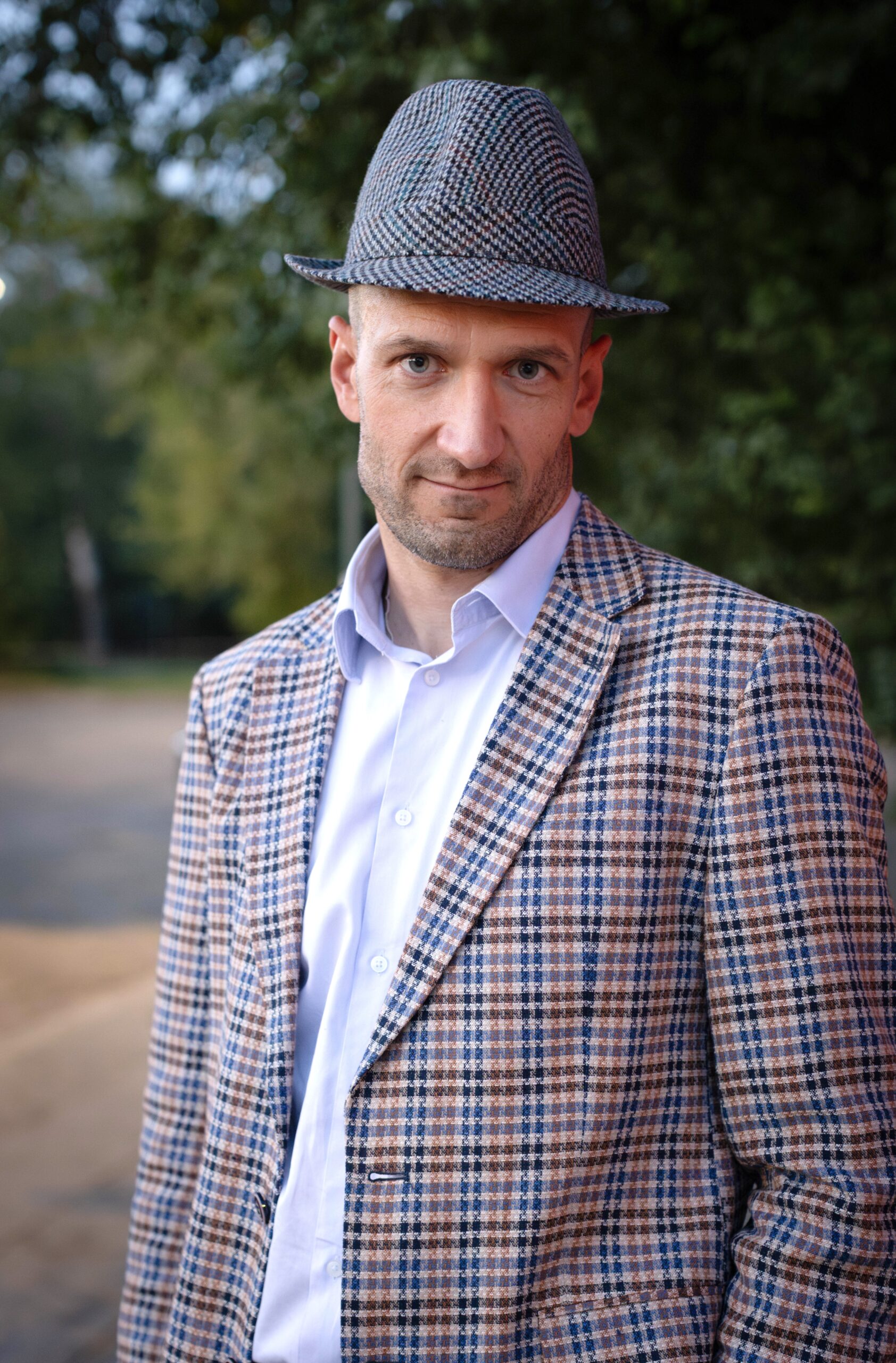 Mężczyzna 45 lat w kraciastej marynarce i kraciastym kapeluszu, z lekkim zarostem, z powagą i lekkim uśmiechem pozuje do zdjęcia wizerunkowego