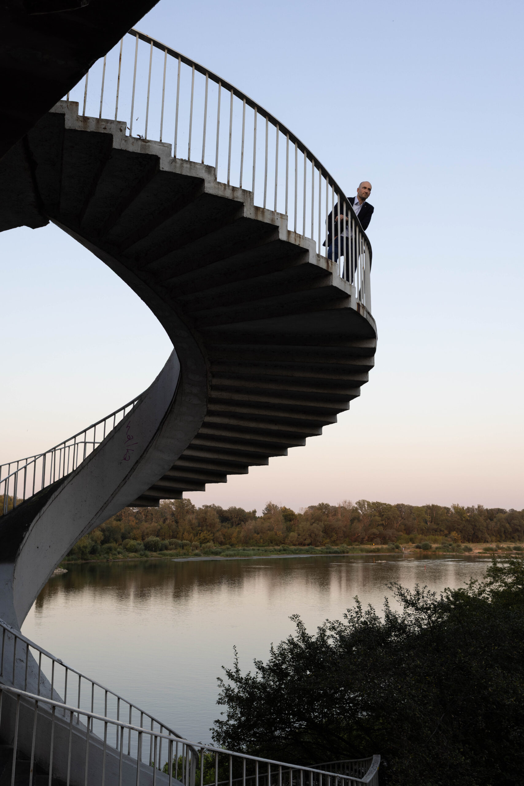 Mężczyzna 45 lat w eleganckim stroju business casual w nietypowej sesji wizerunkowej pozuje na kręconych schodach Mostu Gdańskiego na tle Wisły o zachodzie słońca