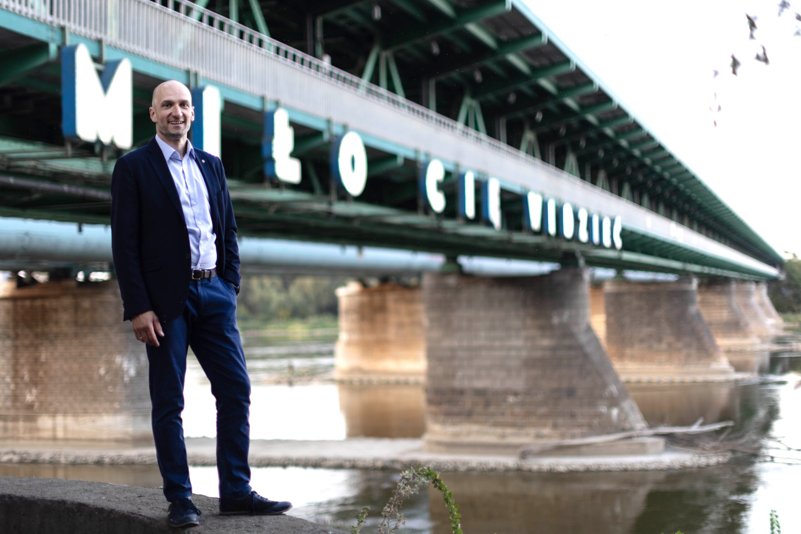 Mężczyzna 45 lat w eleganckim stroju business casual uśmiechnięty pozuje do zdjęcia wizerunkowego na tle Mostu Gdańskiego i neonu Miło Cię widzieć