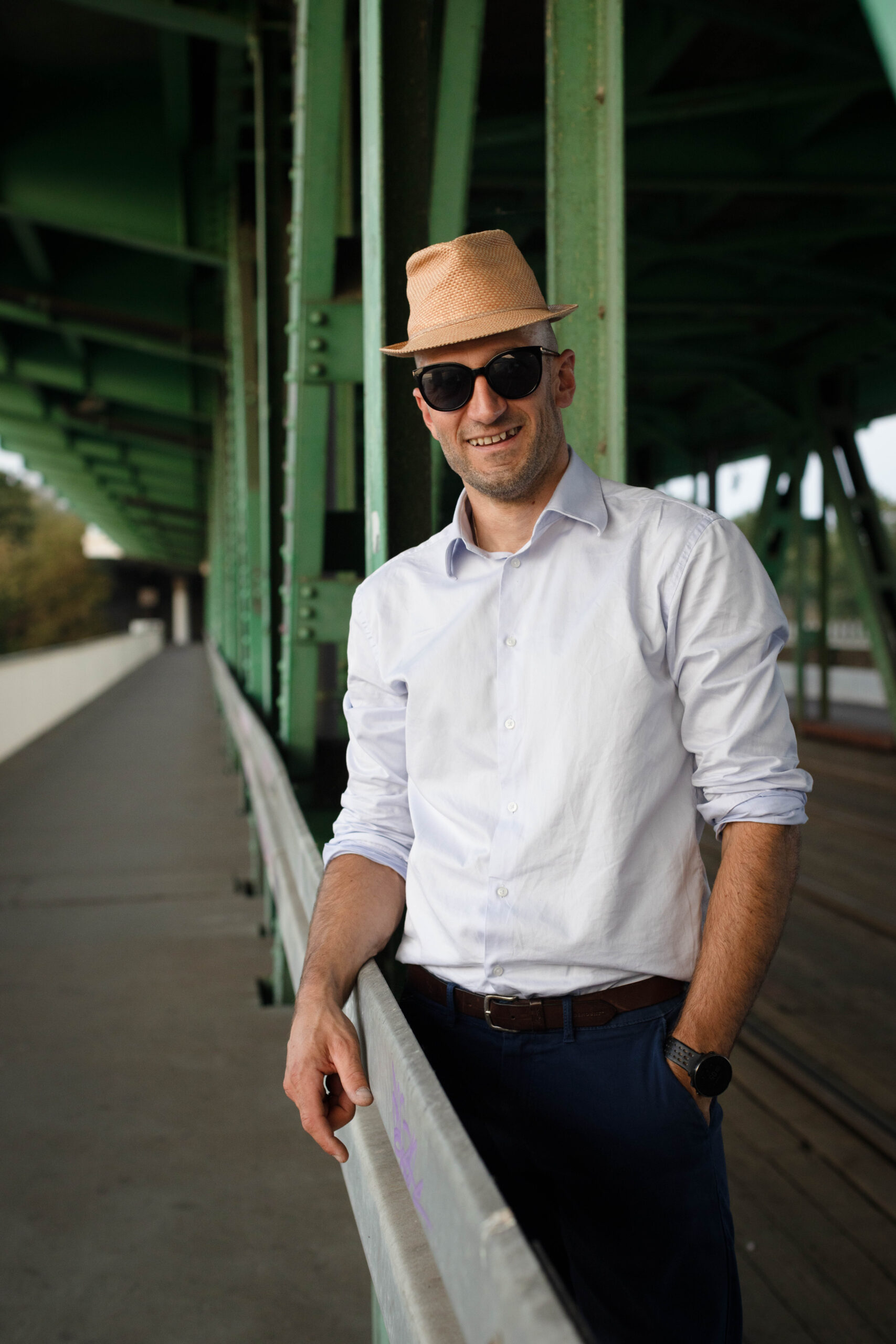 mężczyzna pozuje do zdjęcia na starym moście, w białej koszuli, kapeluszu i ciemnych okularach, z zawadiackim uśmiechem, archetyp Błazen