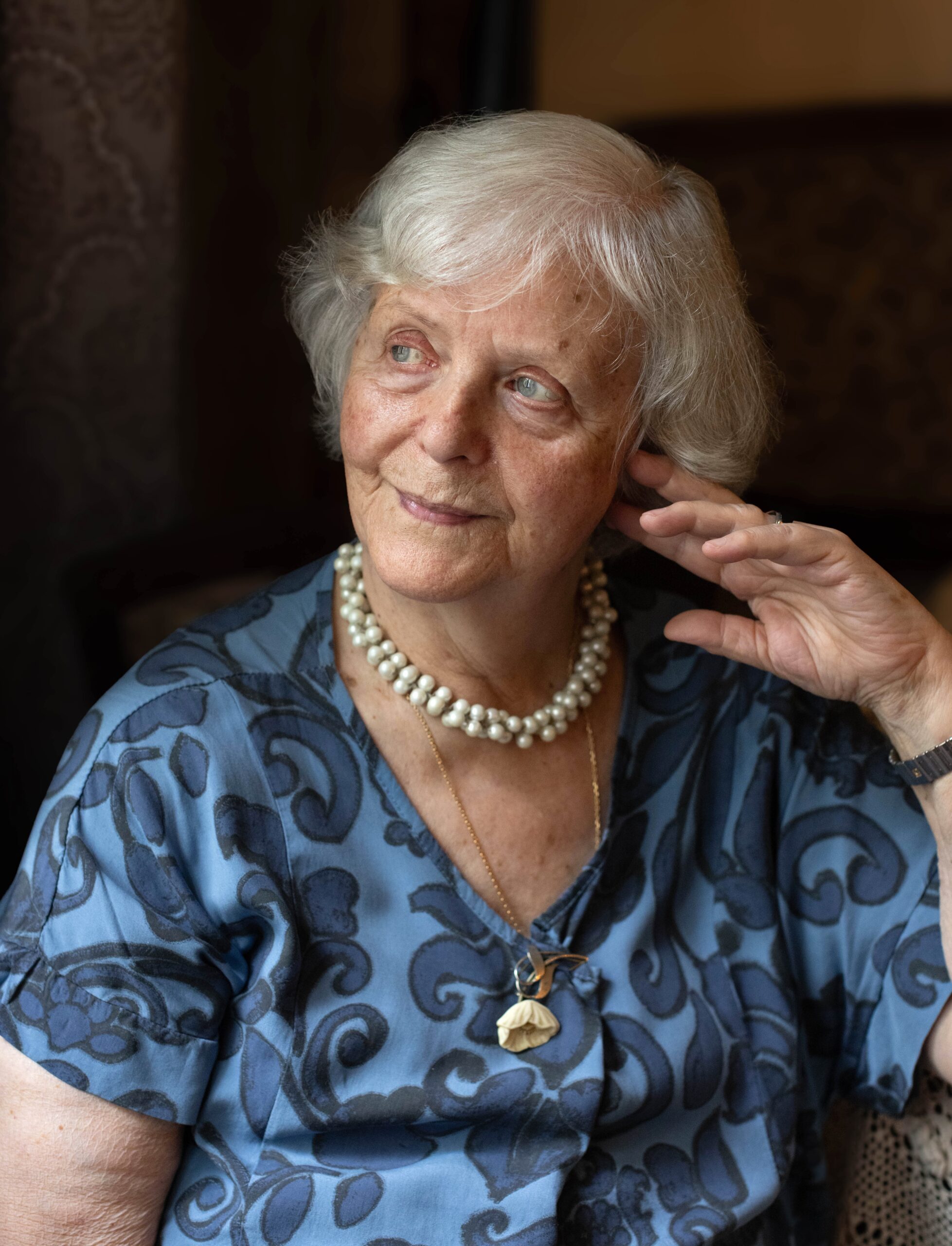 Siwowłosa starsza pani z półuśmiechem patrzy w okno, podpierając się dłonią