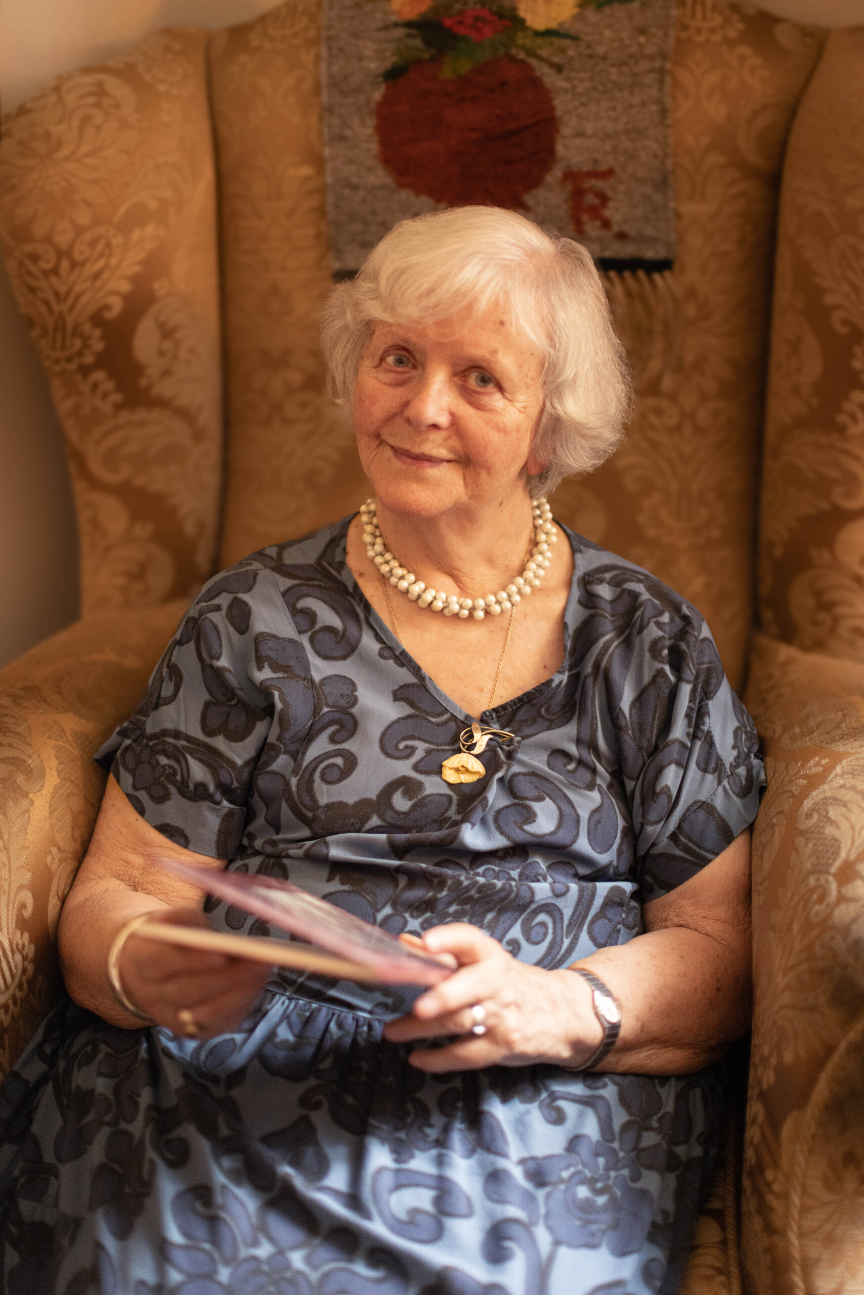 starsza siwa kobieta z uśmiechem patrzy w obiektyw siedząc w fotelu i trzymając w rękach książkę, sesja zdjęciowa dla seniorów prezent na dzień babci i dziadka