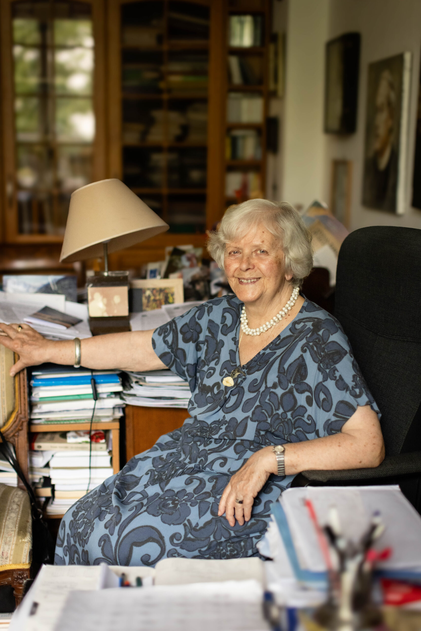 starsza siwa kobieta z uśmiechem patrzy w obiektyw w swoim gabinecie pełnym książek