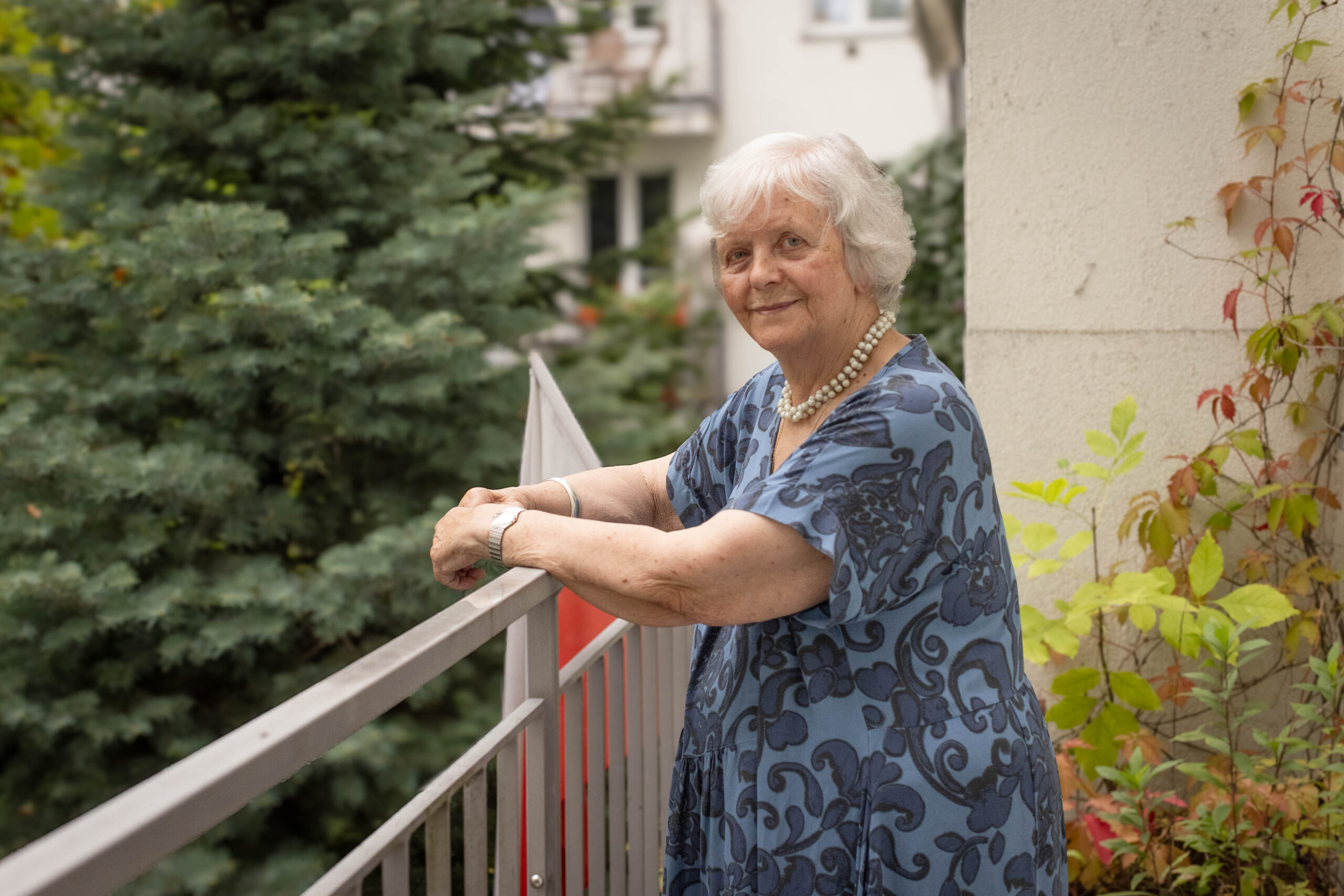 starsza siwa kobieta z uśmiechem patrzy w obiektyw na swoim balkonie, udekorowanym flagą