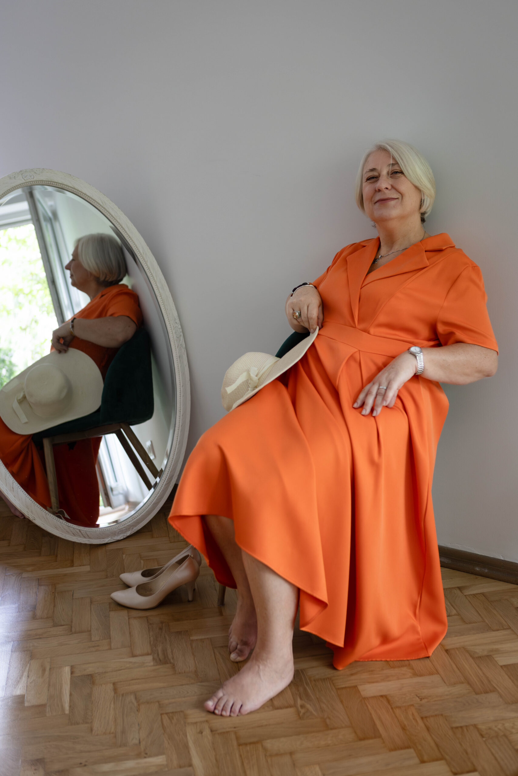 Piękna siwa kobieta w pomarańczowej sukni z uśmiechem pozuje do zdjęcia siedząc na fotelu