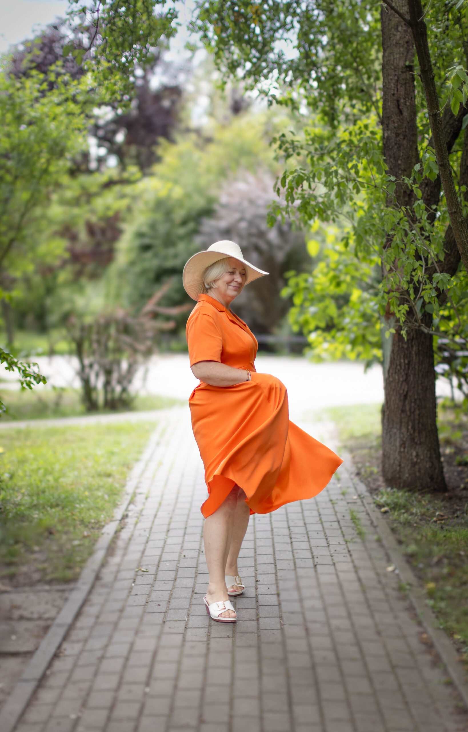 Piękna siwa kobieta w pomarańczowej sukni i jasnym kapeluszu z uśmiechem tańczy na chodniku w parku