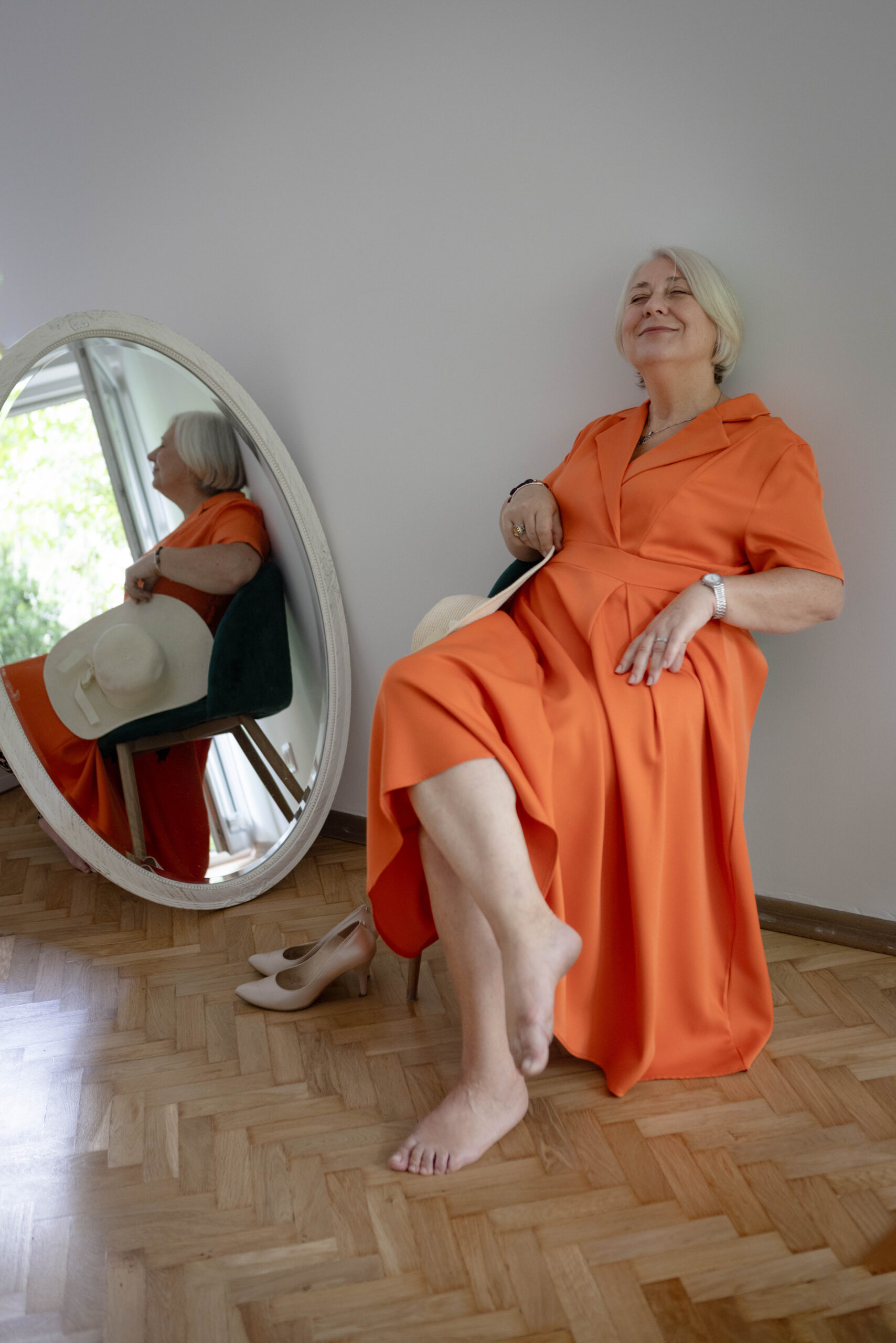 Uśmiechnięta siwowłosa kobieta po sześćdziesiątce, w pomarańczowej sukience i o bosych stopach, która po raz pierwszy przyszła na profesjonalną sesję zdjęciową