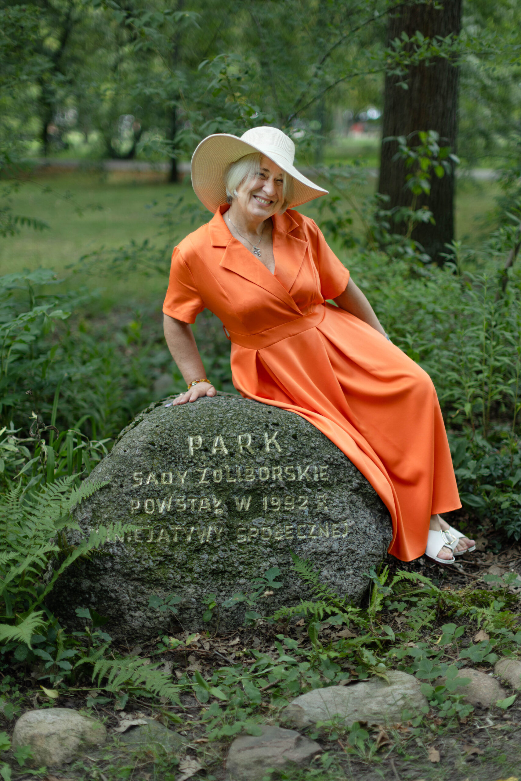 Kobieta sześćdziesięcioletnia w pomarańczowej sukni i jasnym kapeluszu siedzi na kamieniu w parku na sesji zdjęciowej