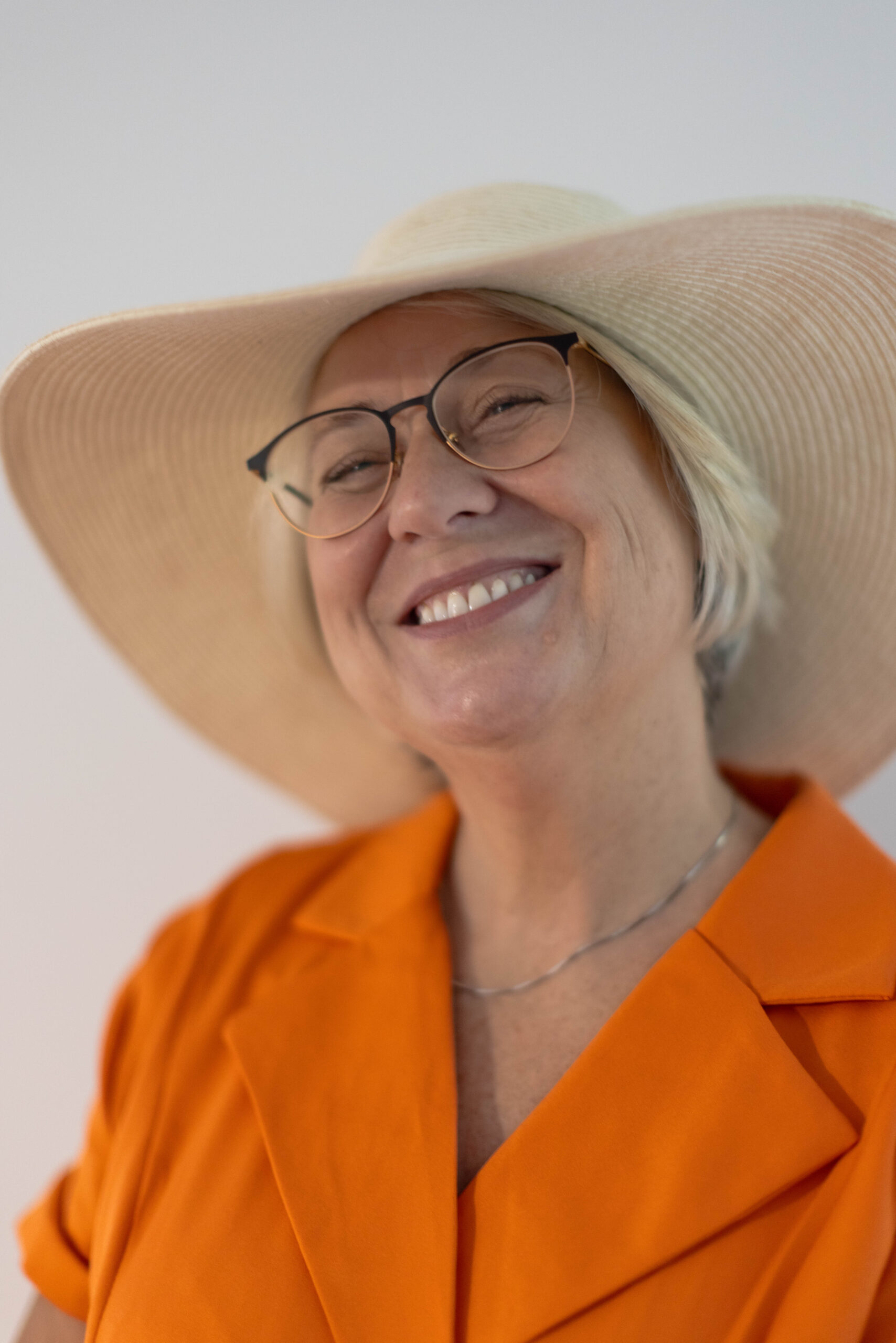 Uśmiechnięta siwowłosa kobieta po sześćdziesiątce, w pomarańczowej sukience i dużym jasnym kapeluszu, która po raz pierwszy przyszła na profesjonalną sesję zdjęciową