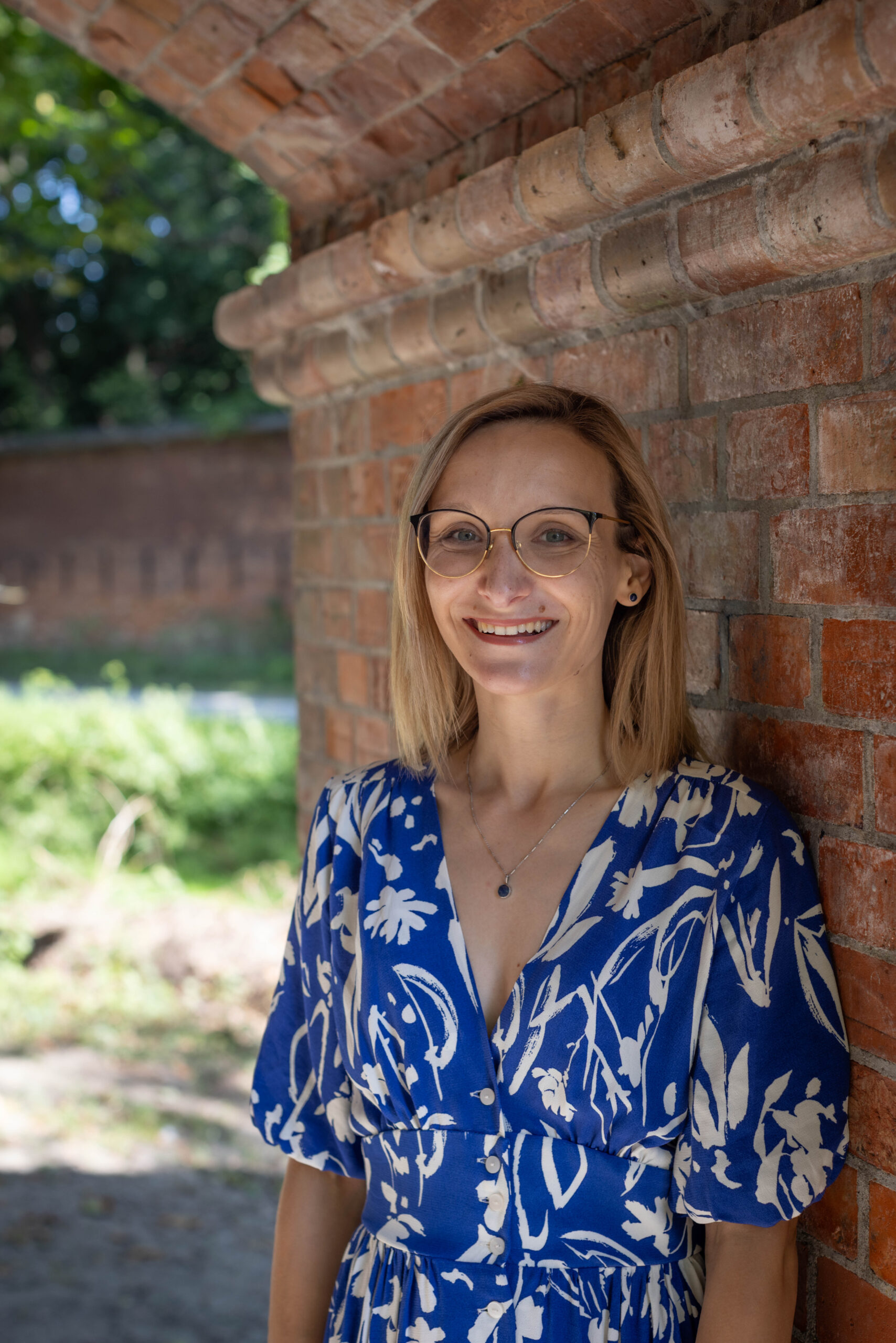 młoda kobieta blondynka w biało-niebieskiej sukience z uśmiechem pozuje do zdjęcia na tle murów ceglanych warszawskiej cytadeli