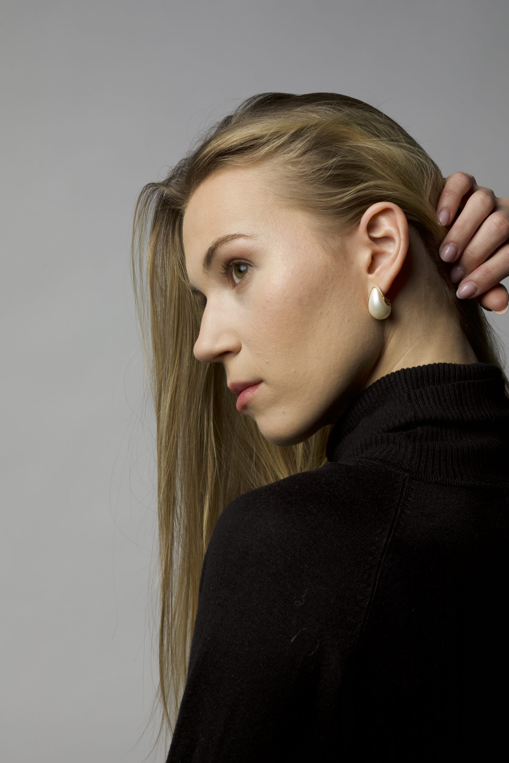 Młoda piękna dziewczyna w czarnym golfie, o długich blond włosach pozuje do zdjęcia zgarniając włosy dłonią i patrząc w dal