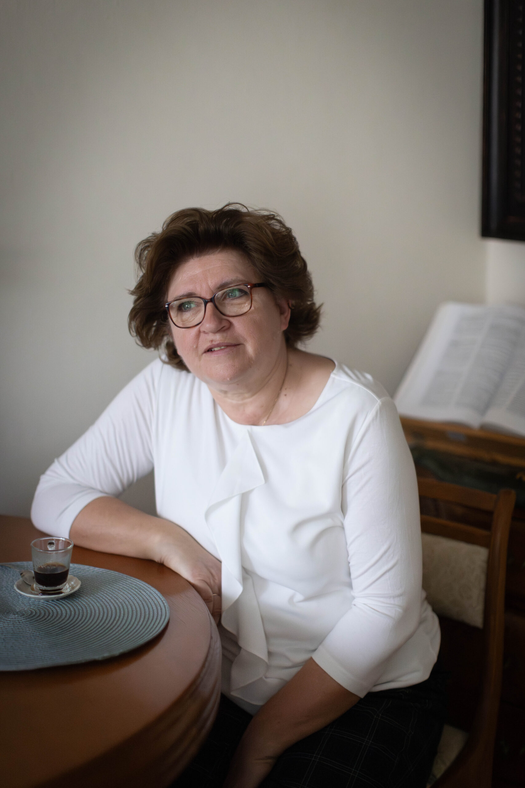 Kobieta koło 60-tki w białej bluzce z rozmarzonym uśmiechem siedzi w domu przy stole, przy filiżance kawy