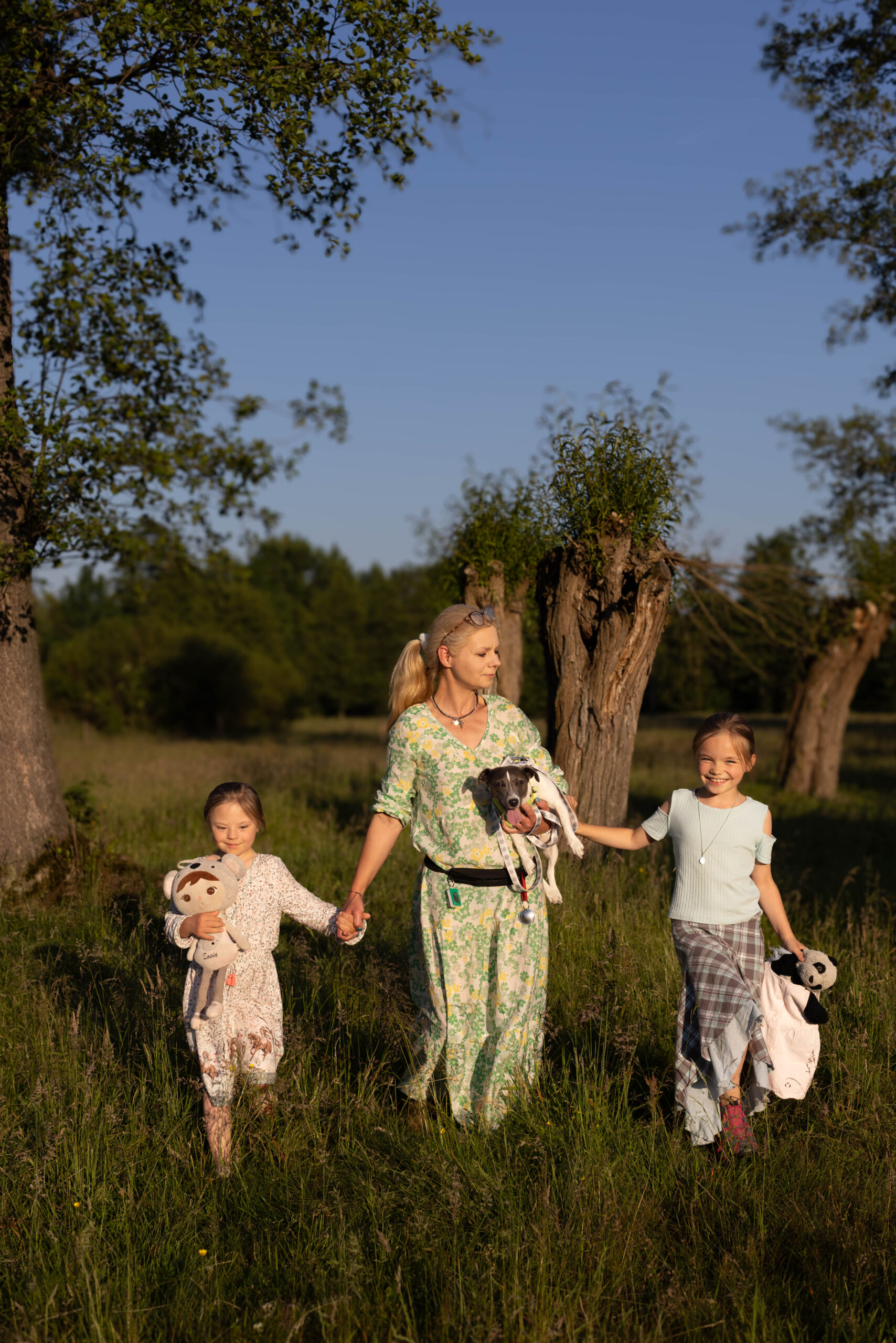 matka z córkami oraz pieskiem idą po łące latem, radosne i uśmiechnięte, w tle las