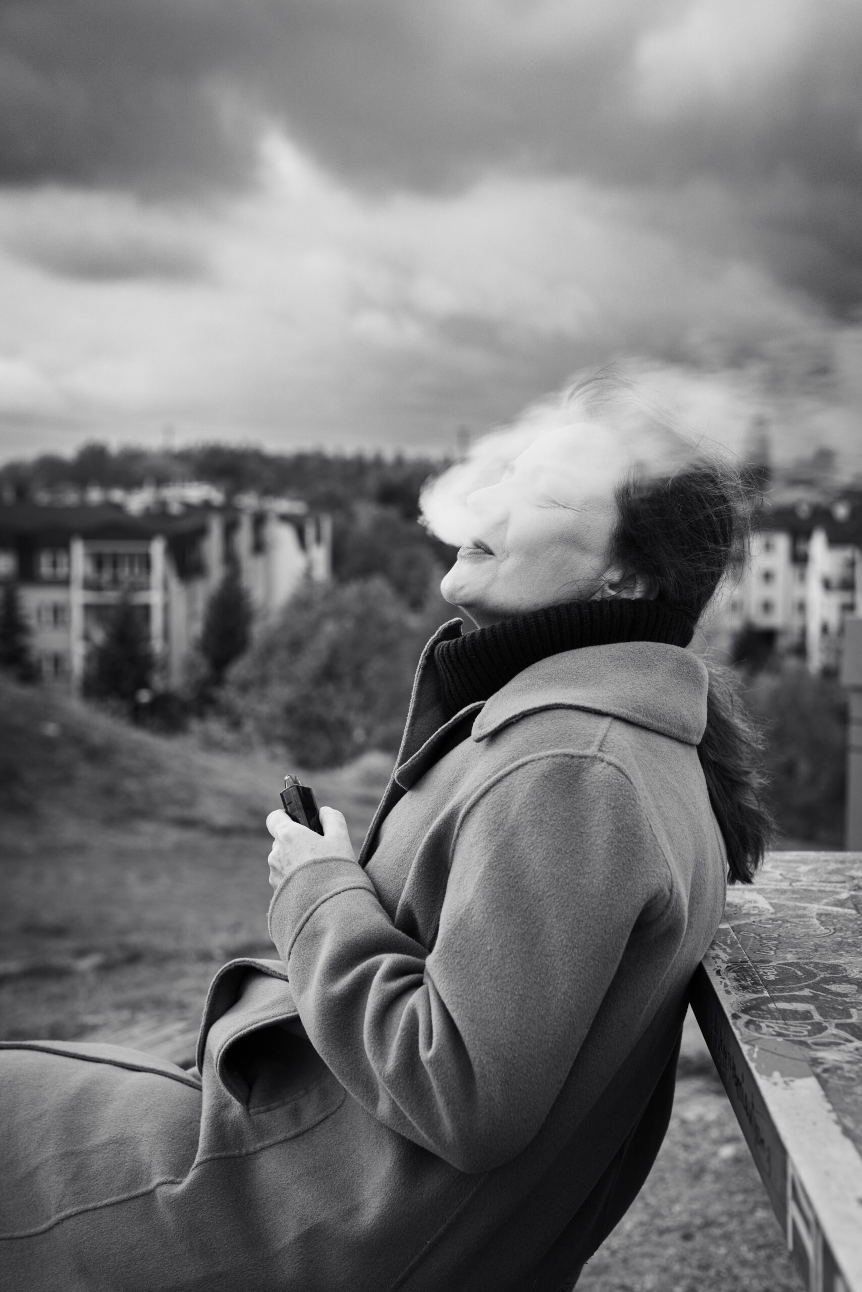 Czarno-białe zdjęcie kobiety na wzgórzu, siedzącej na ławce i puszczającej dym z papierosa