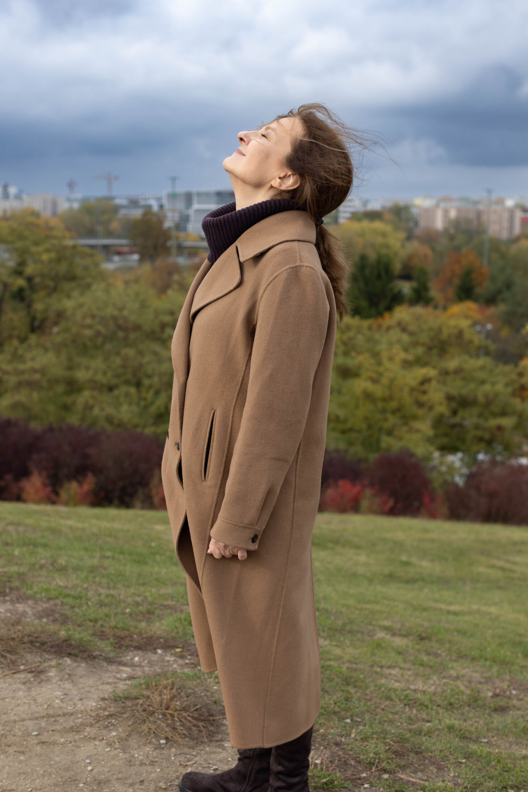 Kobieta stoi na wzgórzu i podstawia twarz pod wiatr, w tle jesienny krajobraz