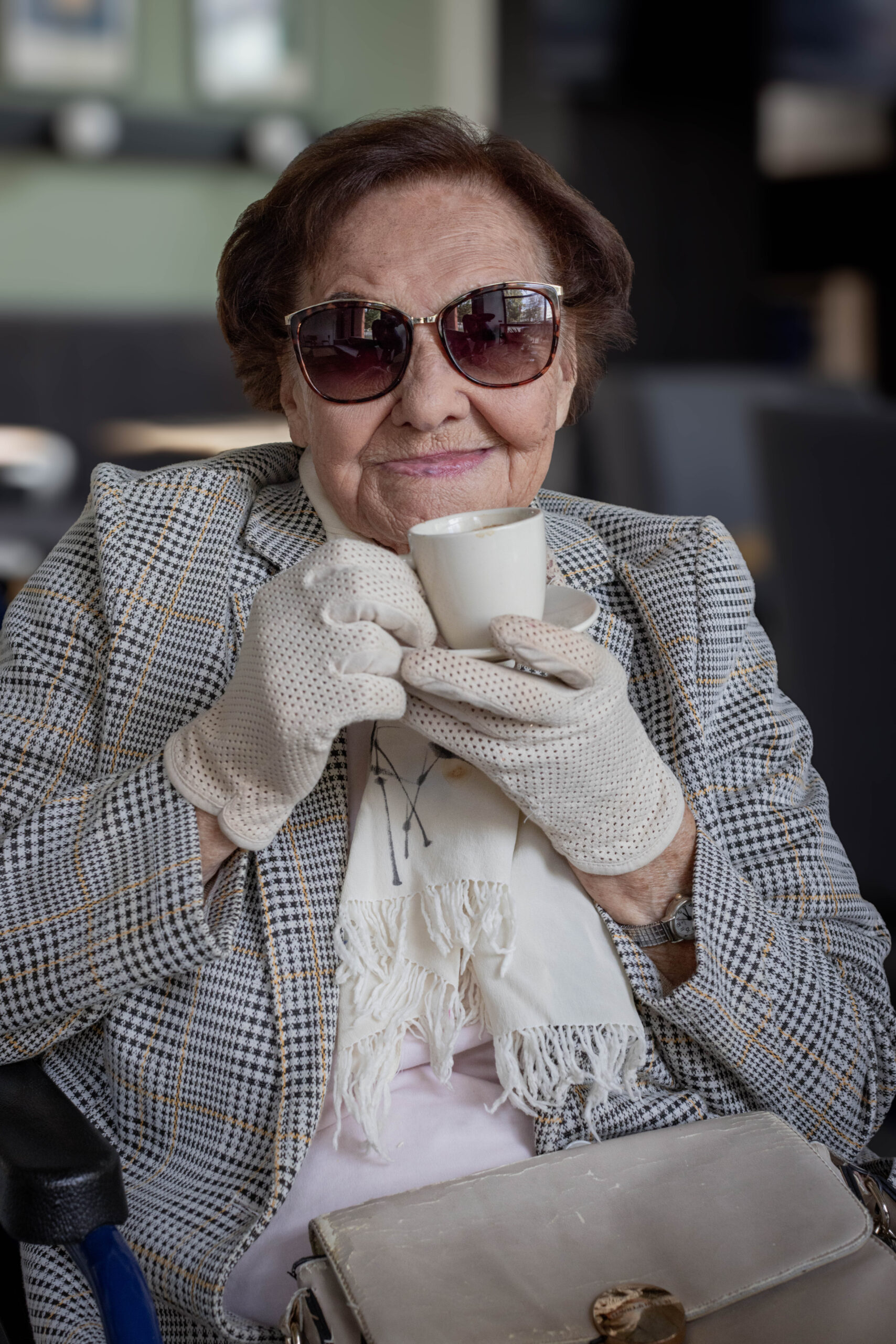 Starsza pani w kawiarni, w marynarce i koronkowych rękawiczkach, z uśmiechem trzyma w dłoni filiżankę kawy