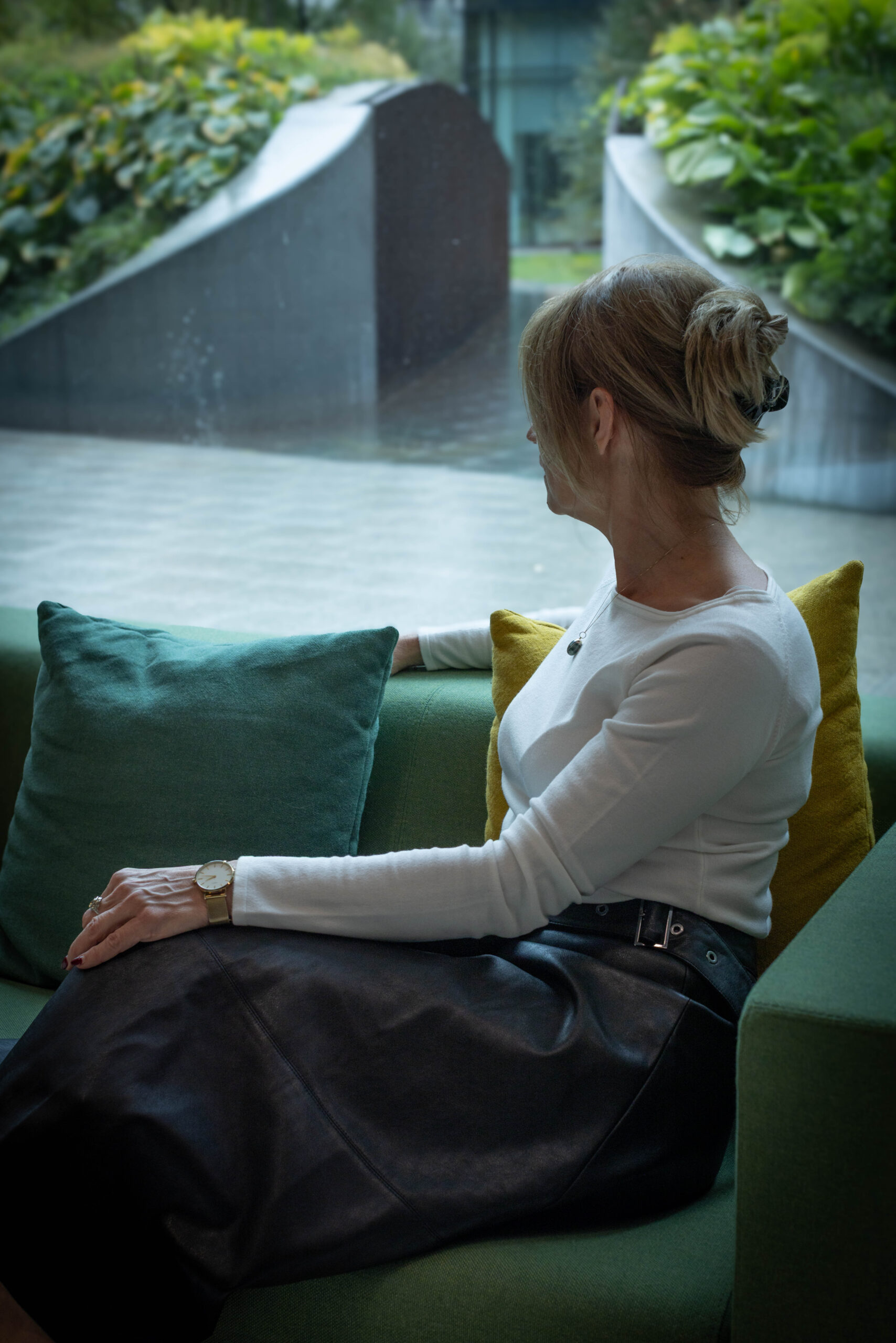 Kobieta o spiętych włosach siedzi na zielonej kanapie bokiem i patrzy przez okno