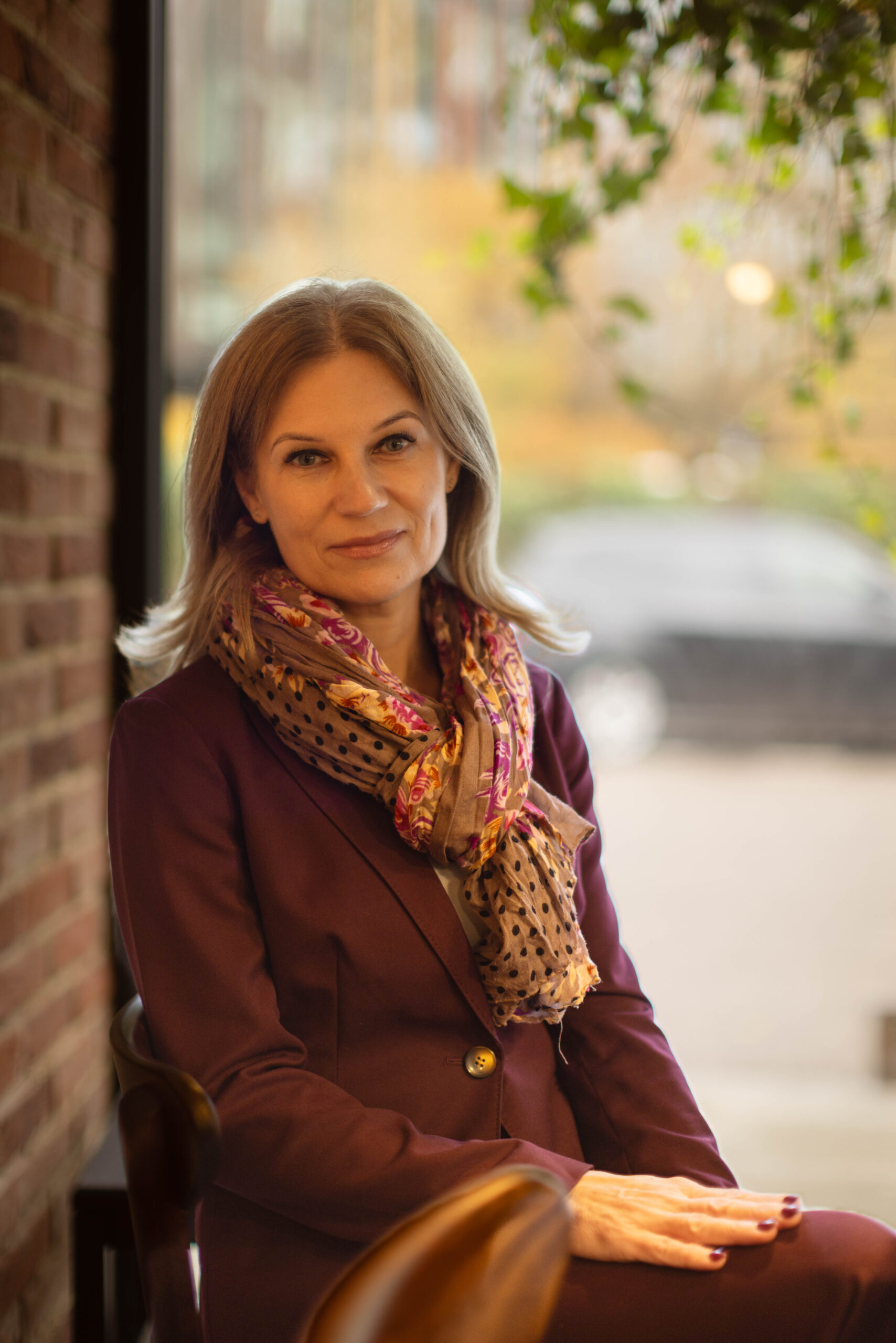 Kobieta w bordowym garniturze pozuje do zdjęcia biznesowego na tle ceglanej ściany i okna