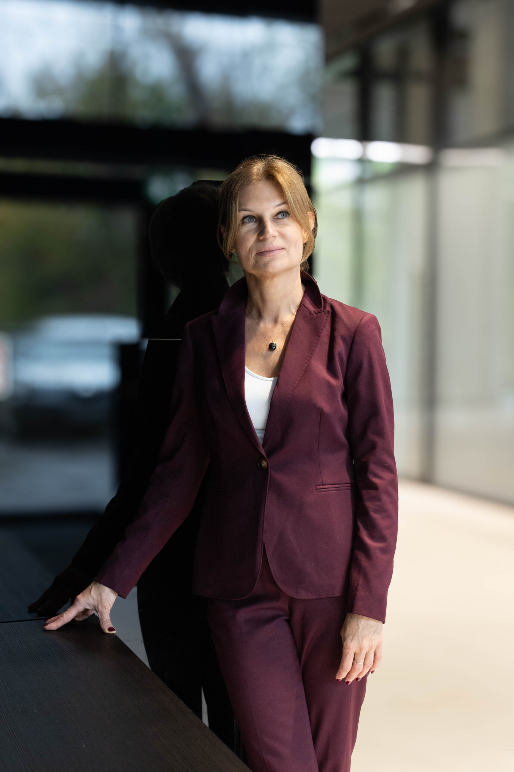 Kobieta w bordowym garniturze pozuje do zdjęcia biznesowego na tle błyszczącej czarnej szyby