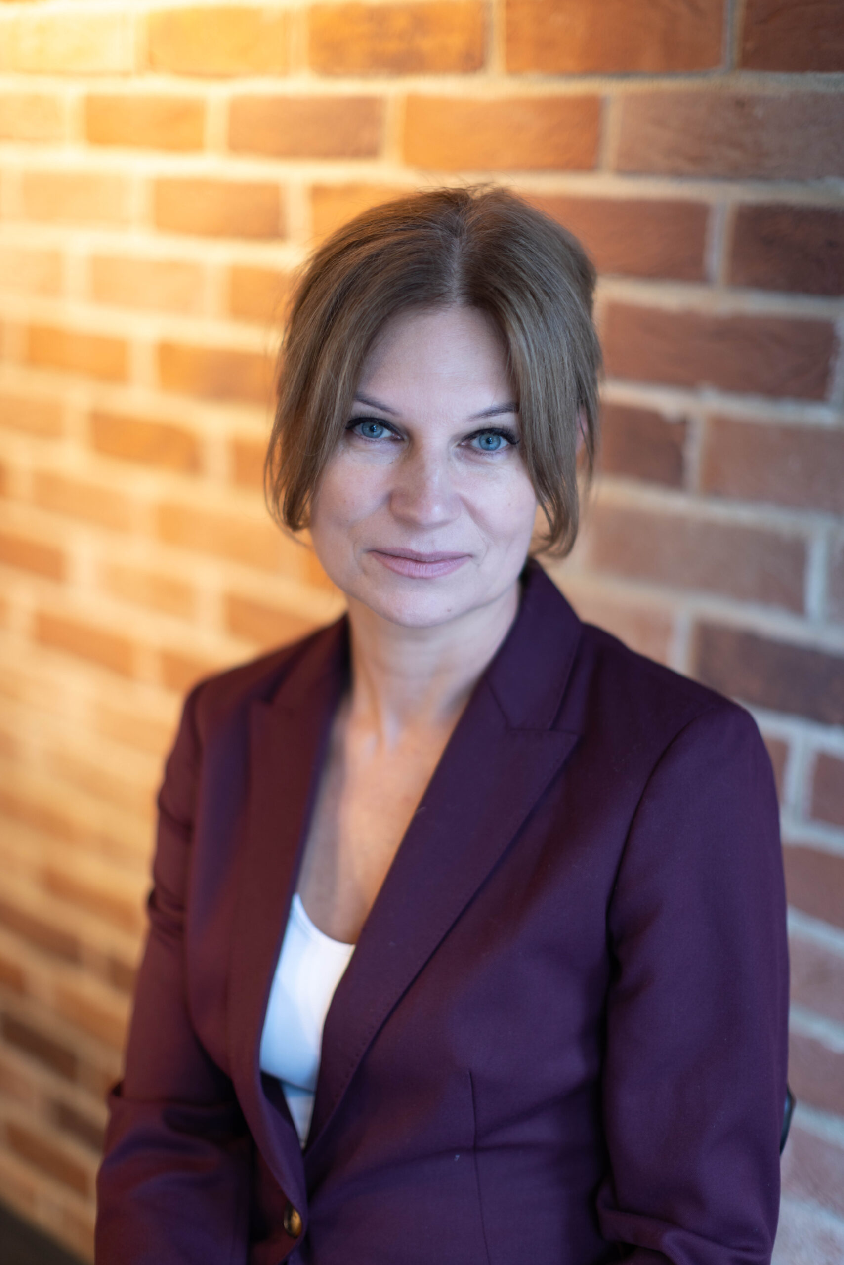 Uśmiechnięta kobieta o spiętych włosach, w bordowym garniturze pozuje do zdjęcia biznesowego na tle ceglanej ściany