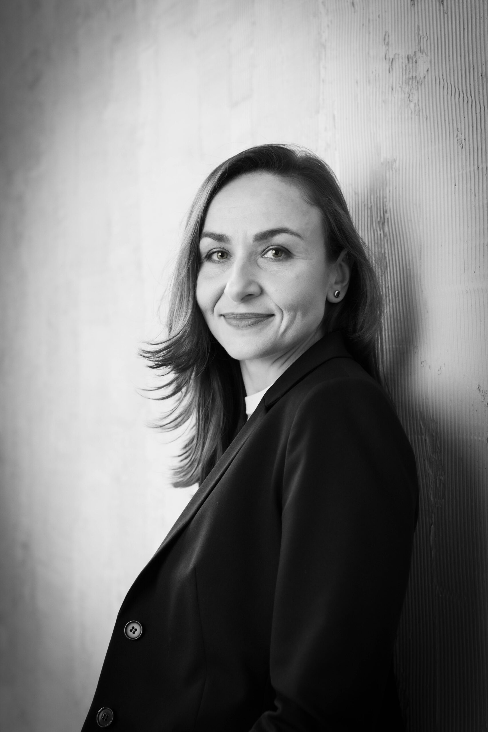 Czarno-białe zdjęcie dziewczyny w marynarce, opartej o ścianę i patrzącej z uśmiechem w obiektyw