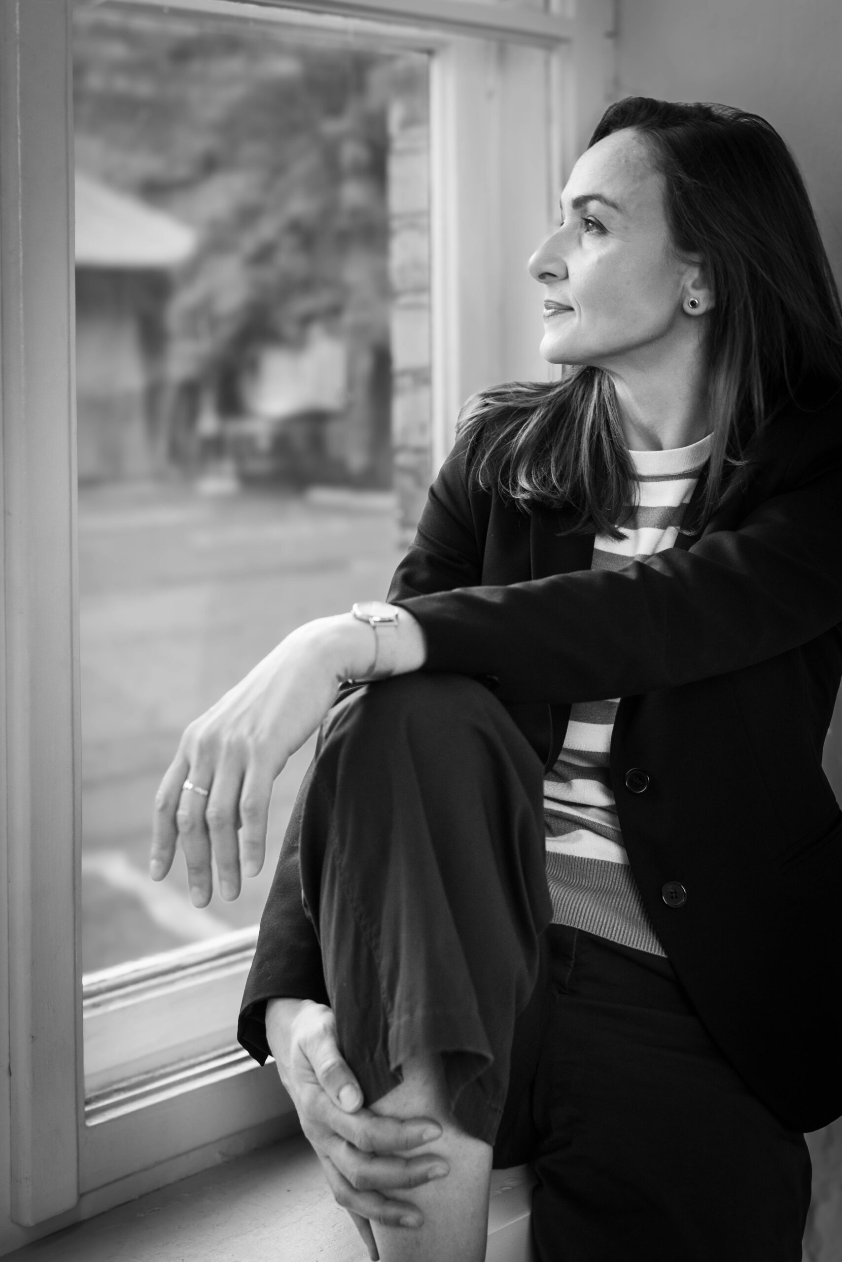 Czarno-białe zdjęcie młodej dziewczyny w marynarce, siedzącej na parapecie i patrzącej przez okno
