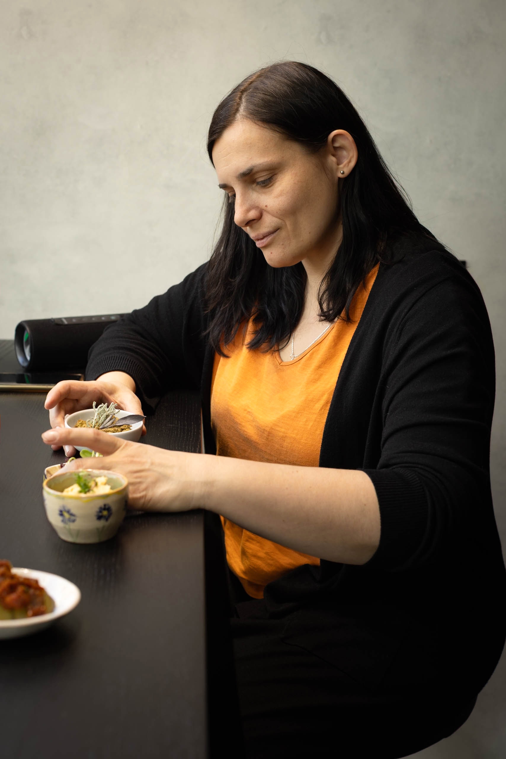 Brunetka w pomarańczowej bluzce i czarnym swetrze układa na ladzie kokilki z pastami udekorowanymi ziołami