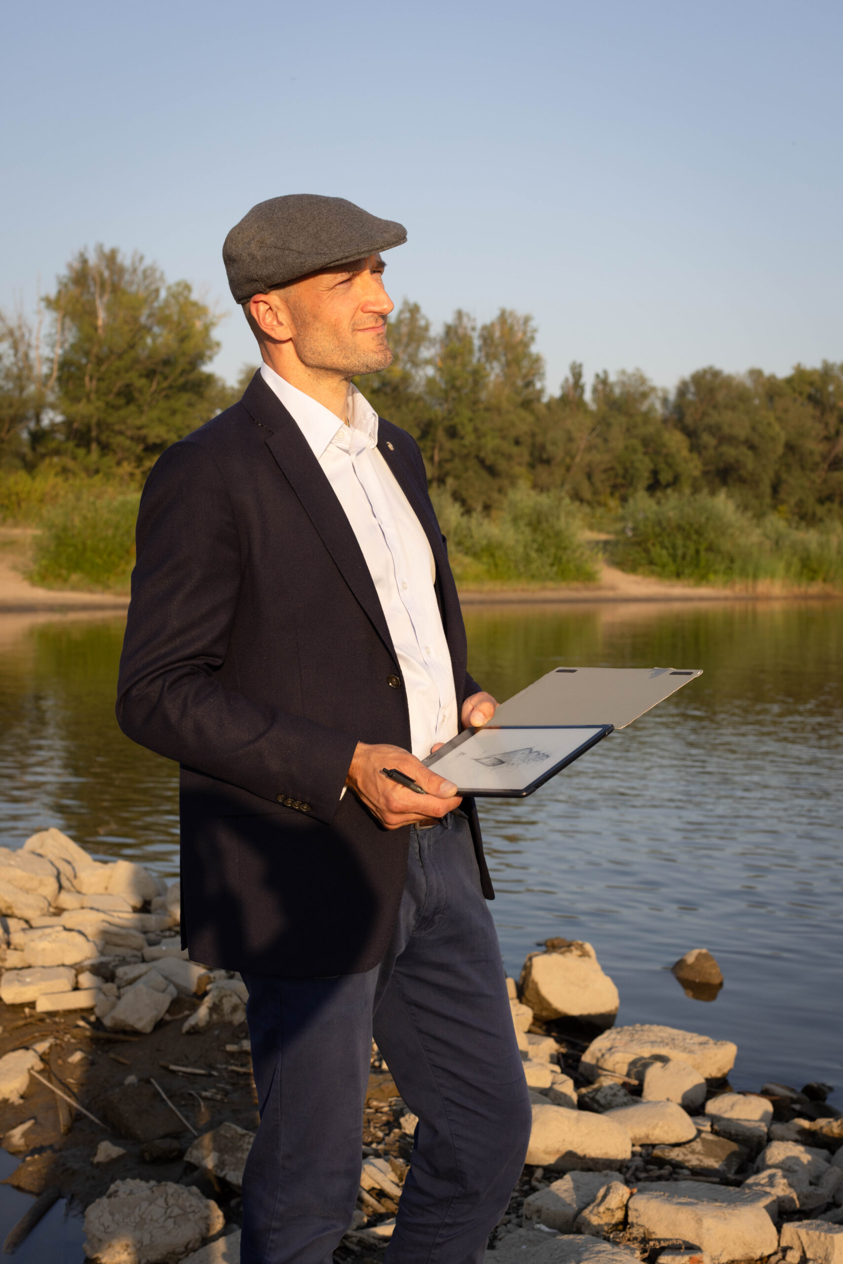 Mężczyzna w marynarce i kaszkiecie pozuje do zdjęcia wizerunkowego na brzegu rzeki, patrząc w kierunku zachodu słońca z tabletem r ręku