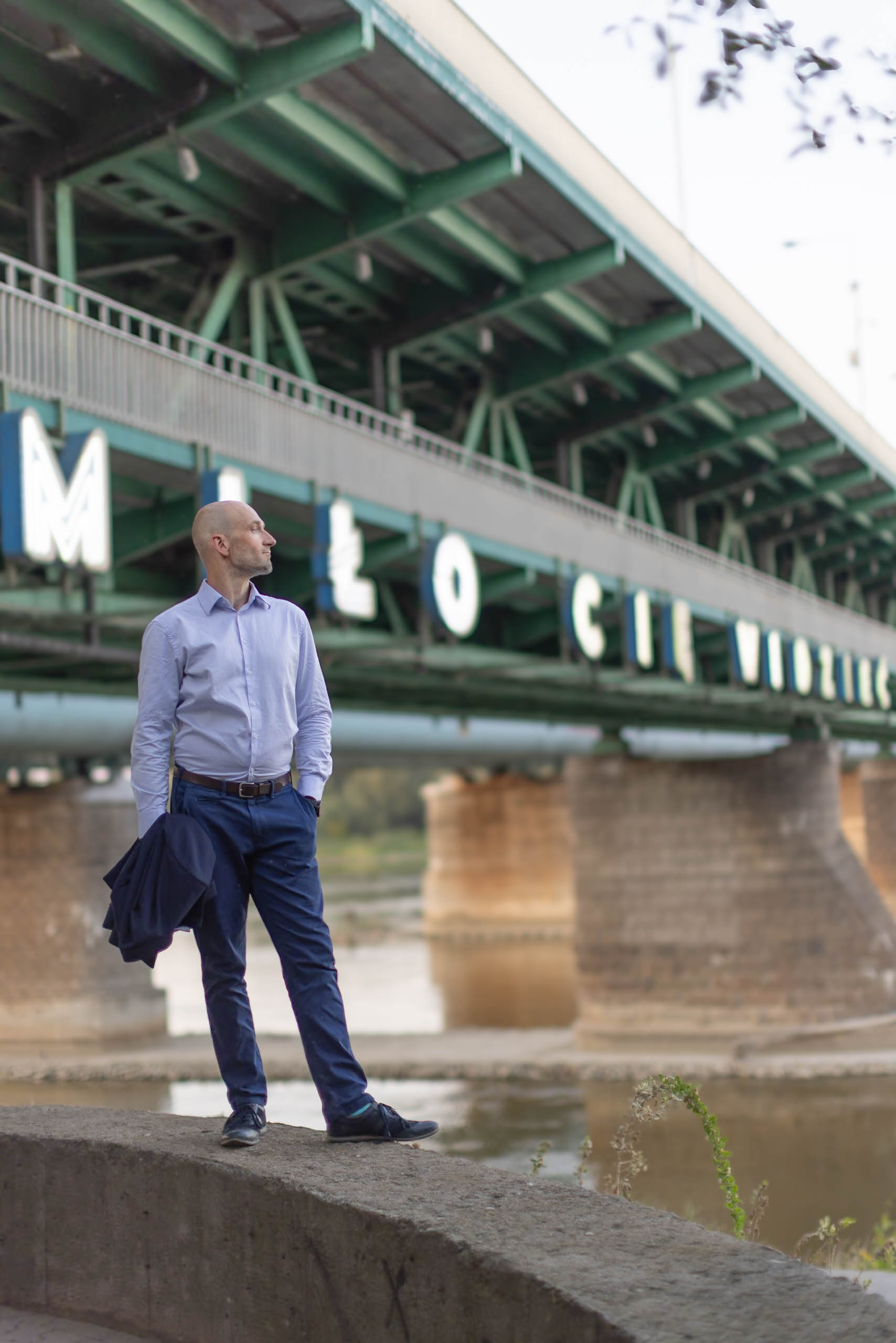 Mężczyzna w koszuli, trzymając w ręku zdjętą marynarkę,pozuje do zdjęcia wizerunkowego na tle mostu i neonu "miło Cie widzieć"