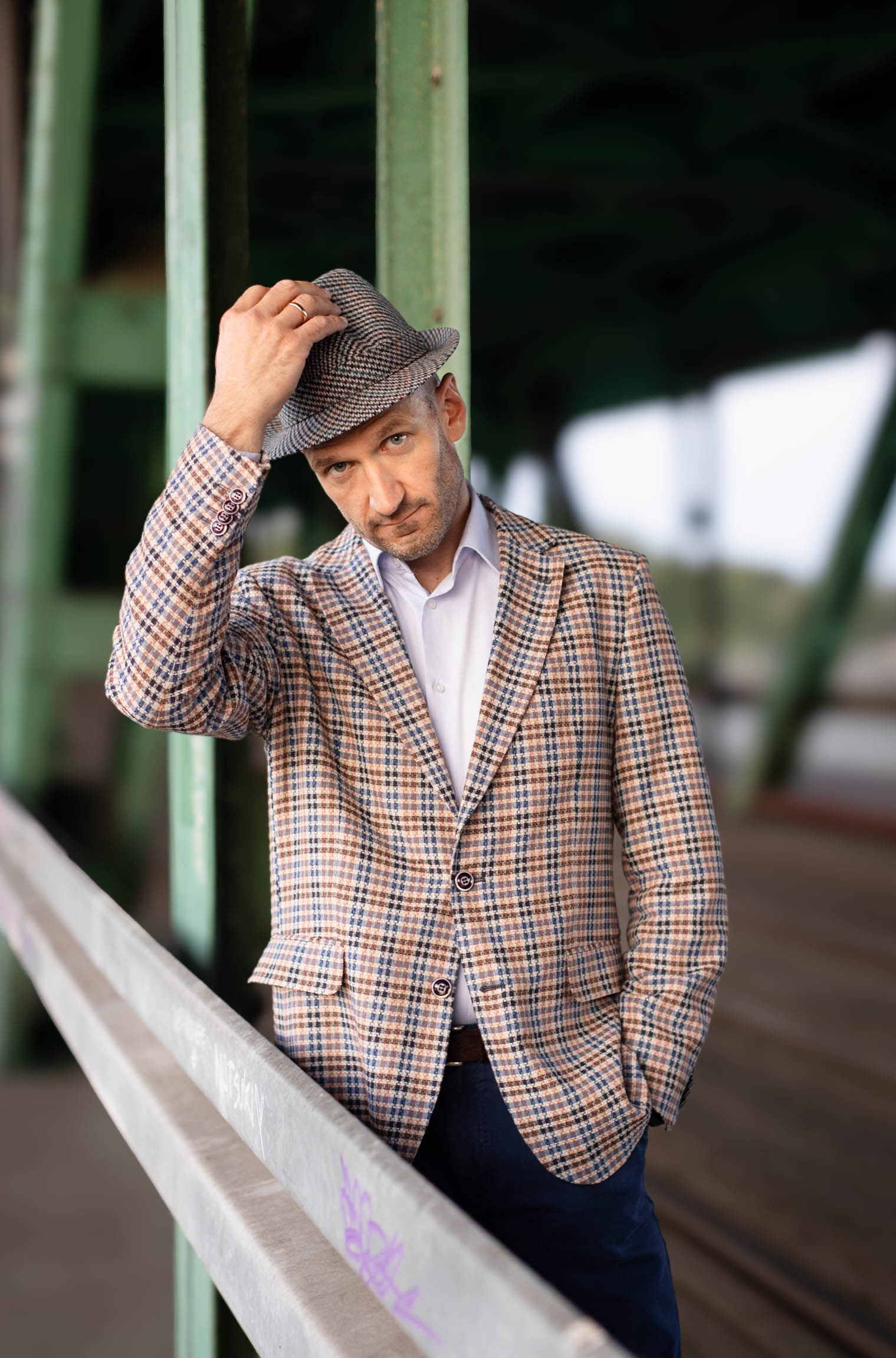 Mężczyzna w marynarce i kapeluszu pozuje do zdjęcia wizerunkowego na moście, uchylając kapelusza