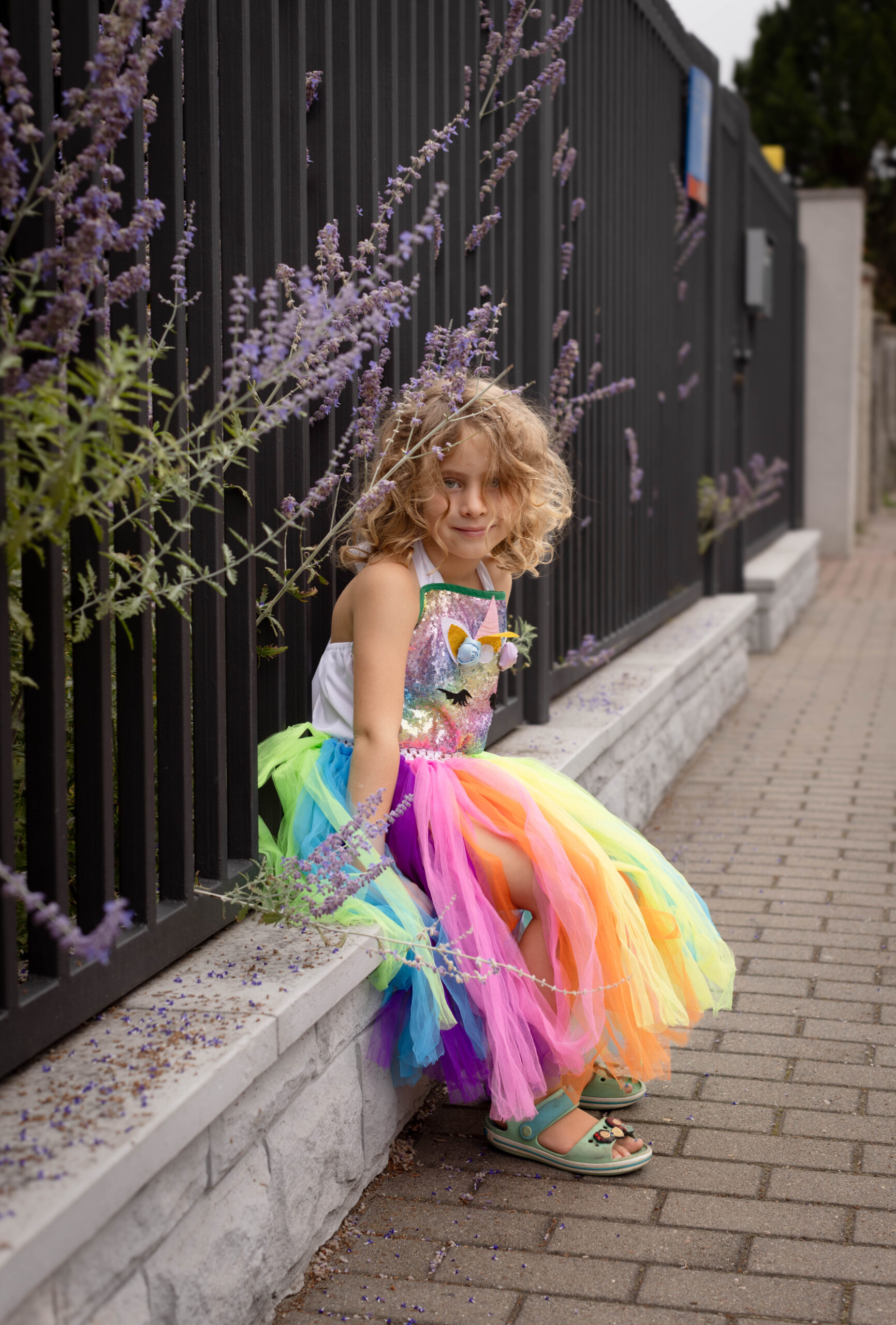dziewczynka w kolorowej tiulowej sukience siedzi na oparciu ogrodzenia i z uśmiechem patrzy w obiektyw