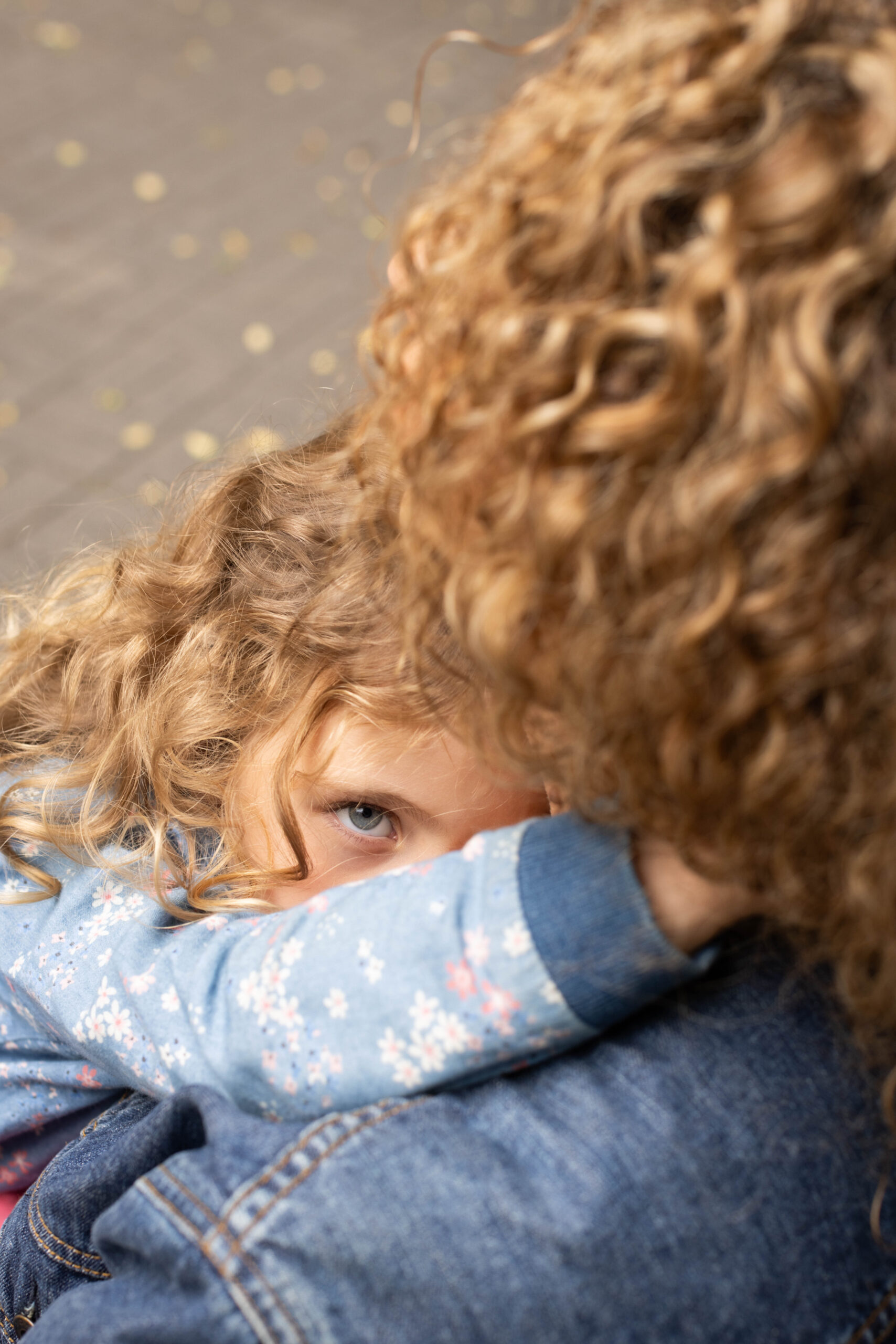 Portret dziewczynki o kręconych jasnych włosach wtulonej w mamę, tak że widać tylko jedno oko dziewczynki