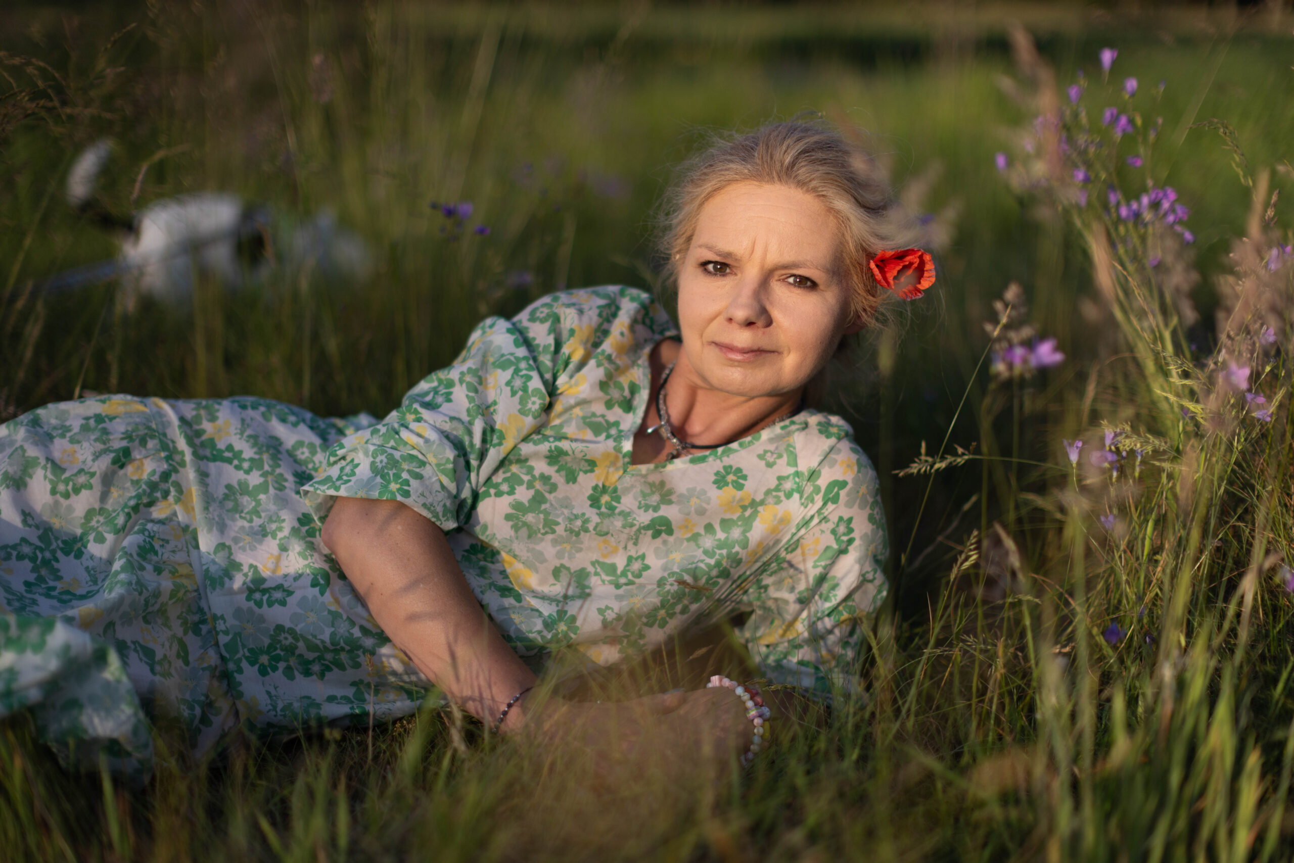 Kobieta w kwiecistej sukience, z makiem we włosach, leży na zielonej trawie wśród kwiatów