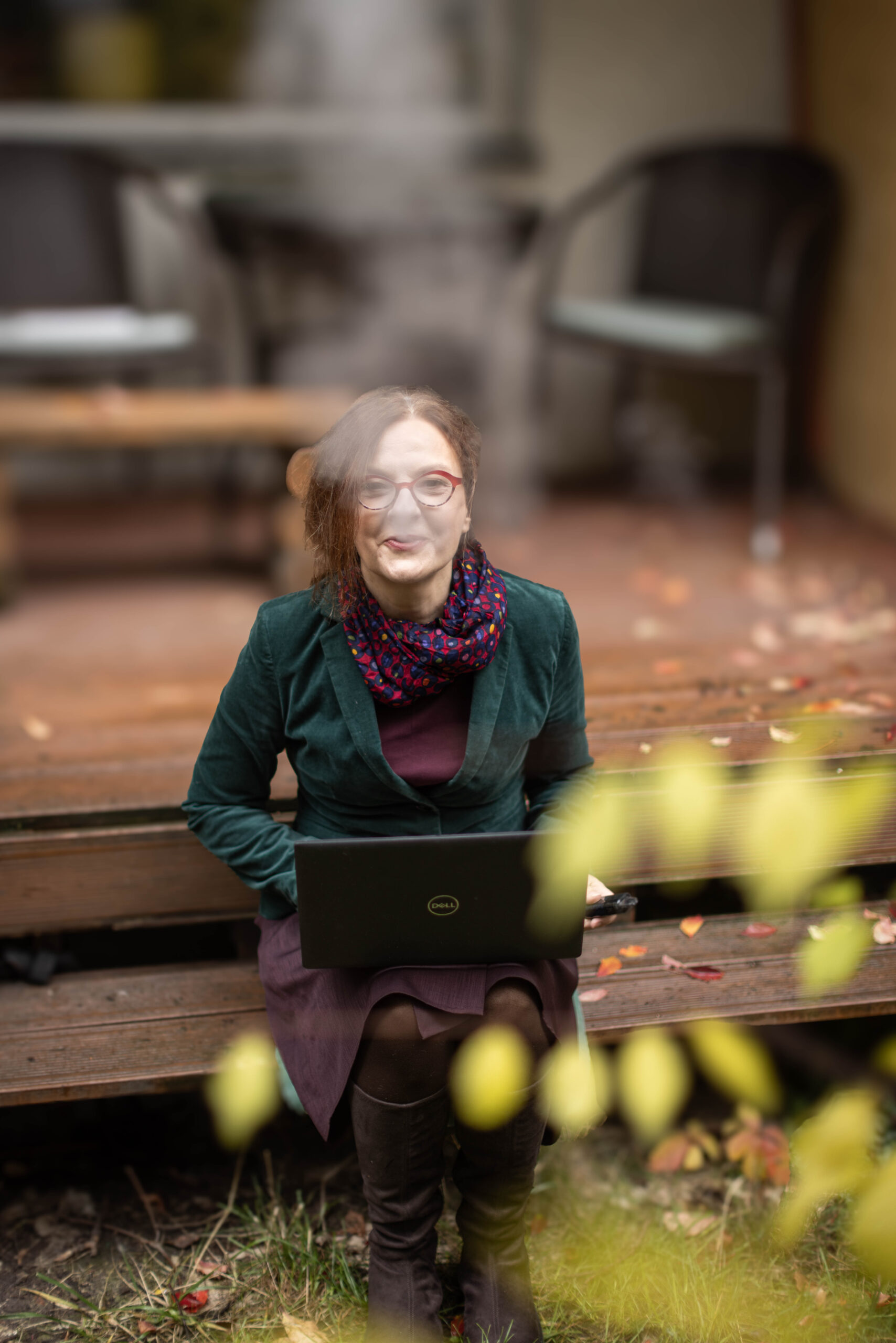 Rudowłosa kobieta w jesiennym ogródku siedzi na schodach z komputerem i puszcza dym z papierosa