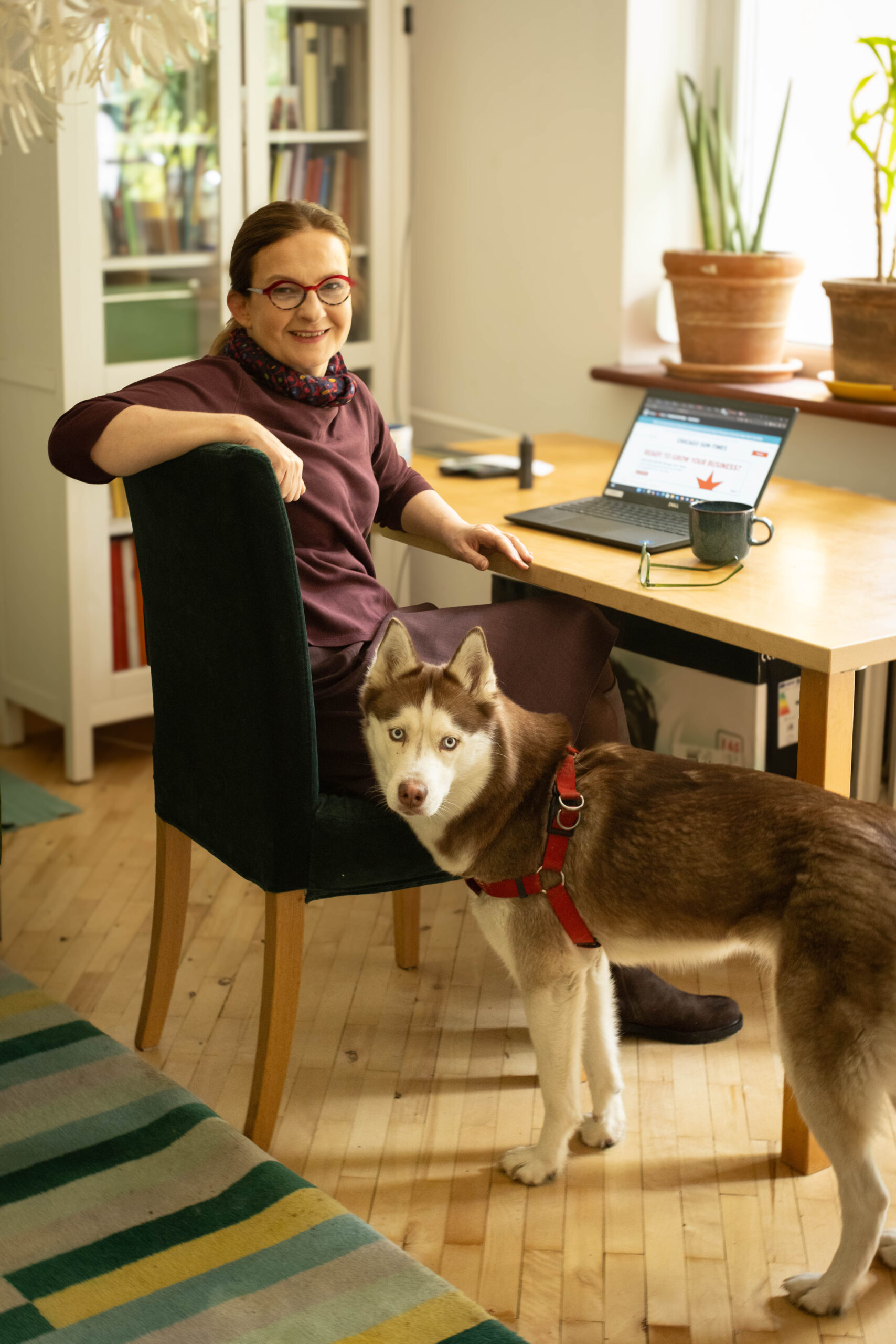 Rudowłosa kobieta w czerwonych okularach siedzi przy biurku, a obok stoi pies husky