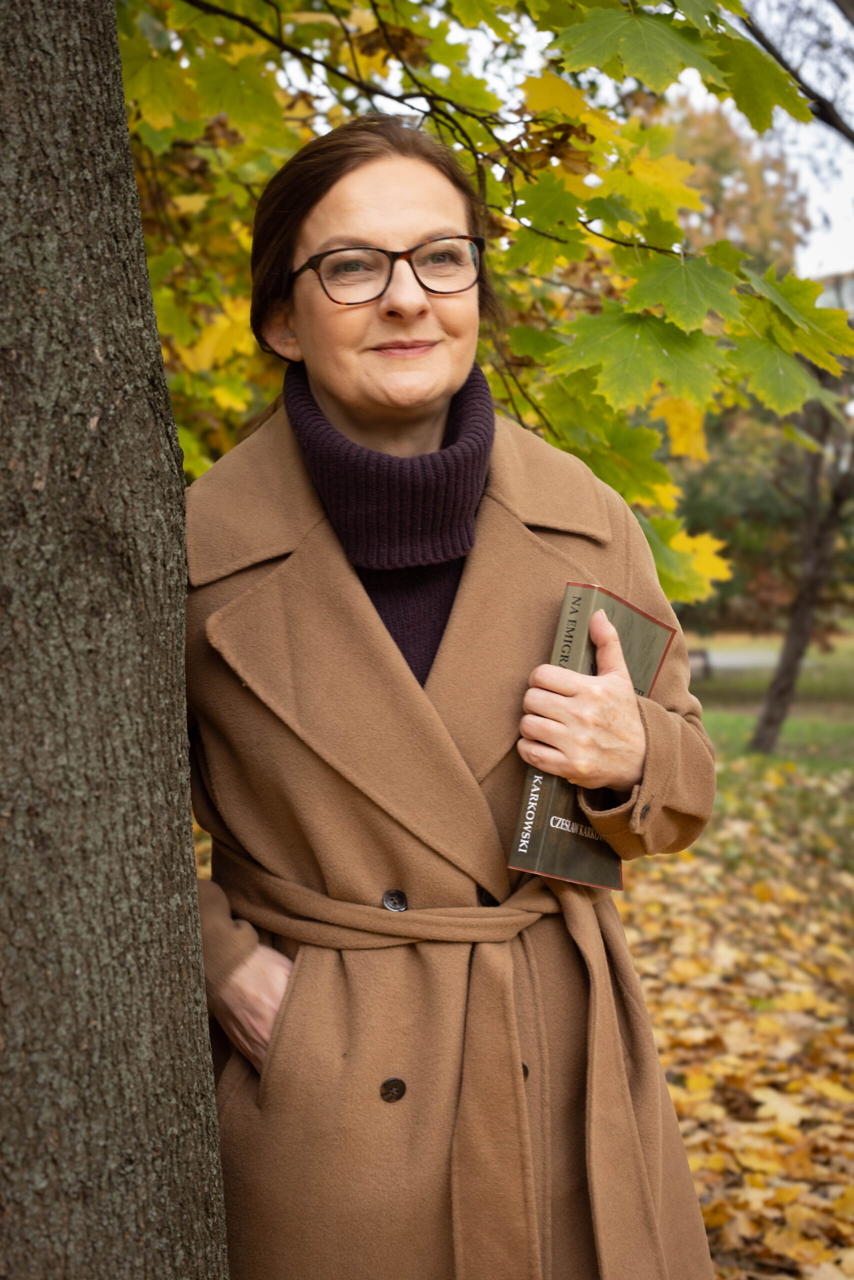 Rudowłosa kobieta w płaszczu camel stoi z książką w parku, oparta o drzewo