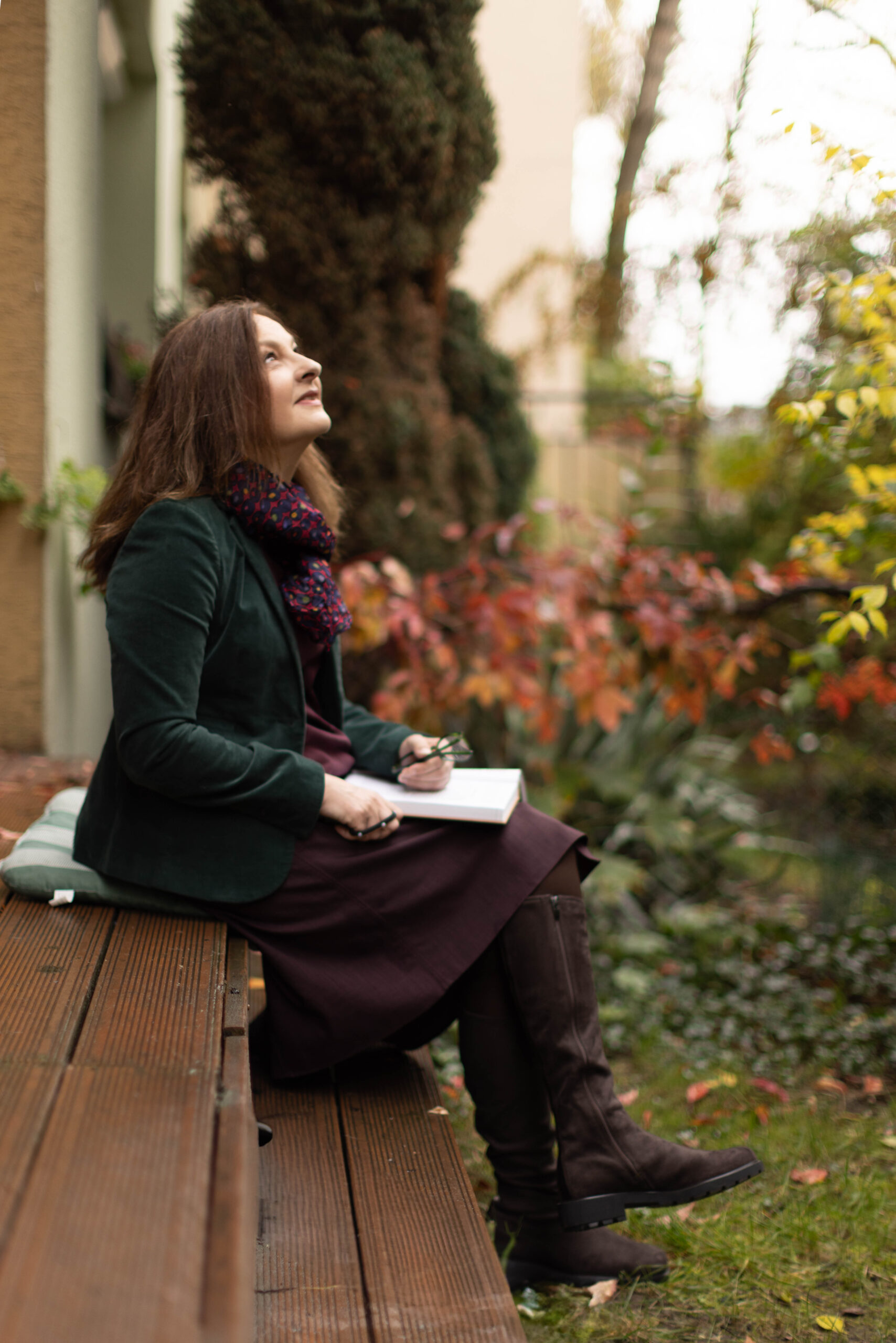 Rudowłosa kobieta siedzi na drewnianych schodach w jesiennym ogródku z książką w rękach
