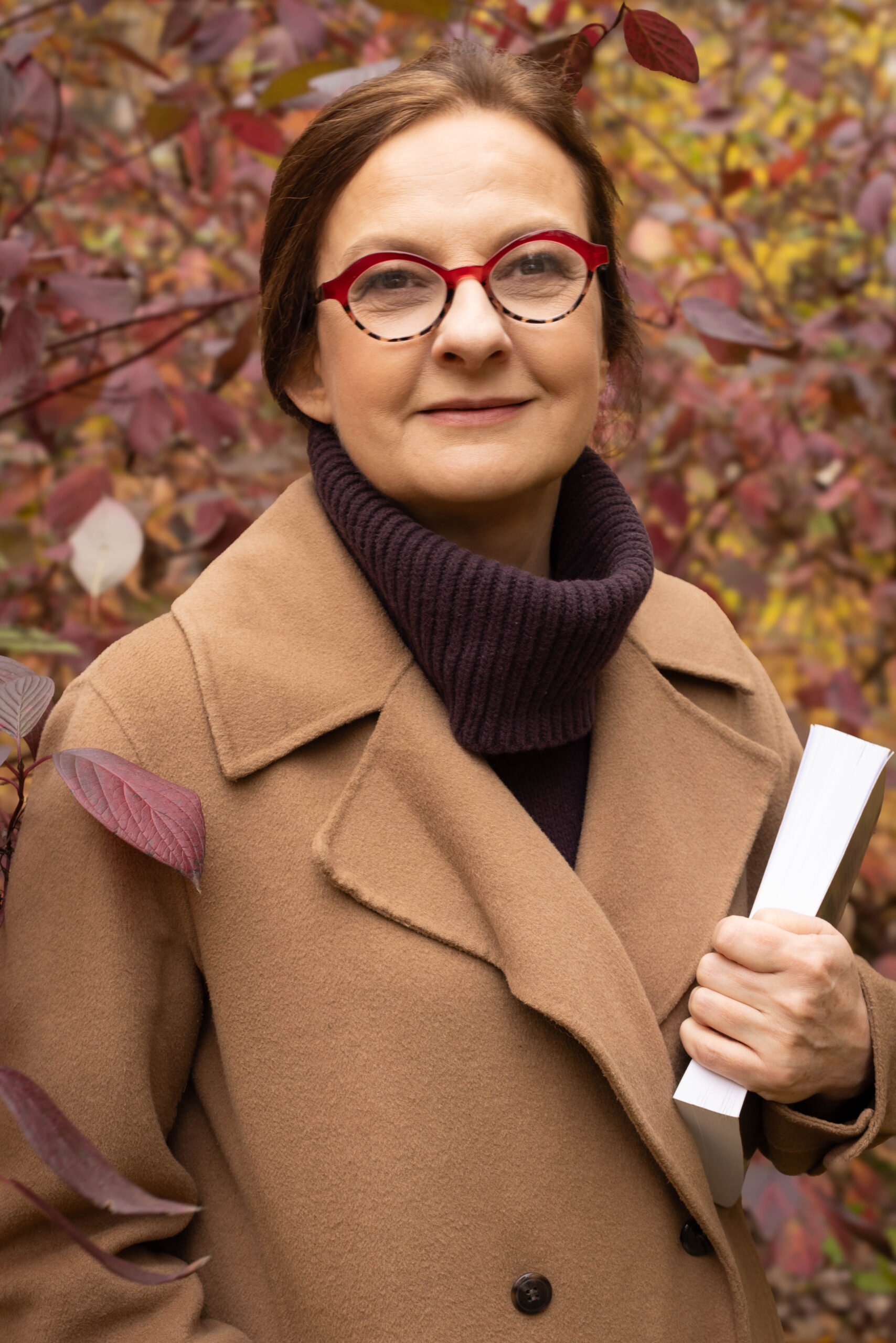 Rudowłosa kobieta w płaszczu camel stoi z książką w parku, na tle kolorowych liści